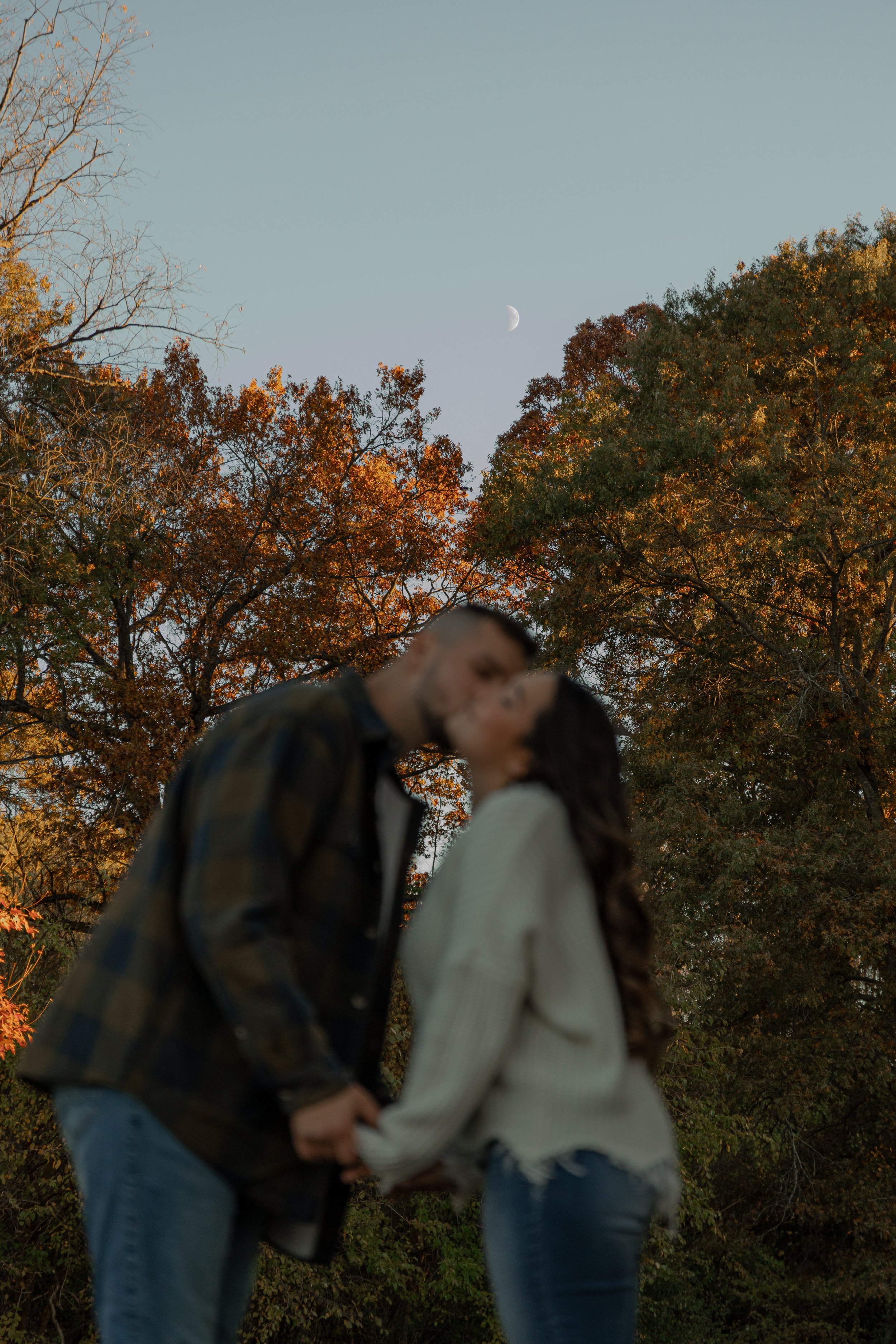A couple holding hands and kissing outdoors during autumn, with orange and green trees and a visible waxing crescent moon in the sky.