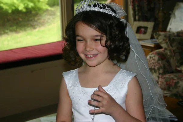 A young girl wearing a white dress, a tiara, and a veil, smiling indoors with a window and a living room in the background.