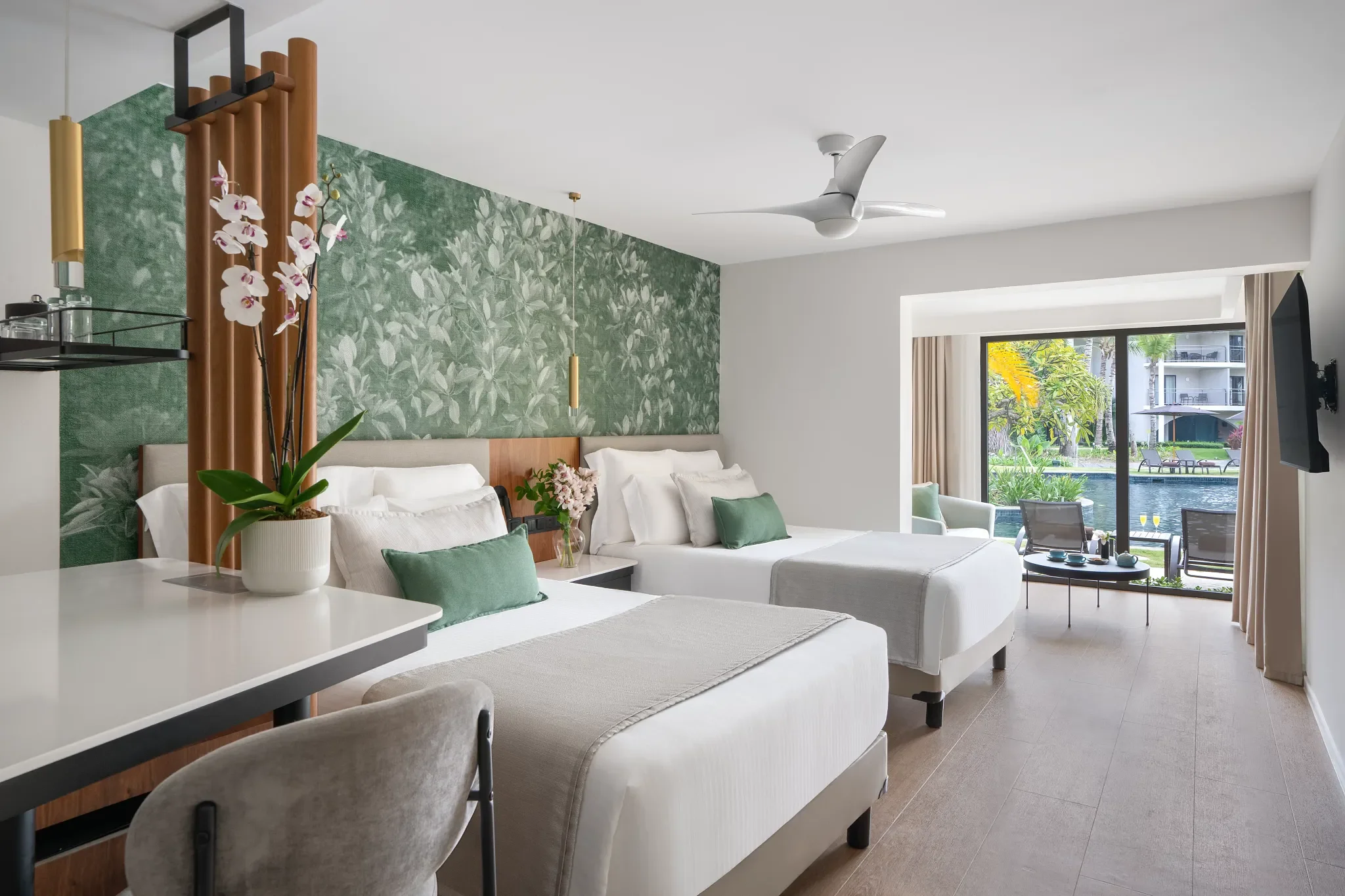Room with two beds, green pillows, floral wall art, balcony with outdoor seating, tropical scenery, and TV.