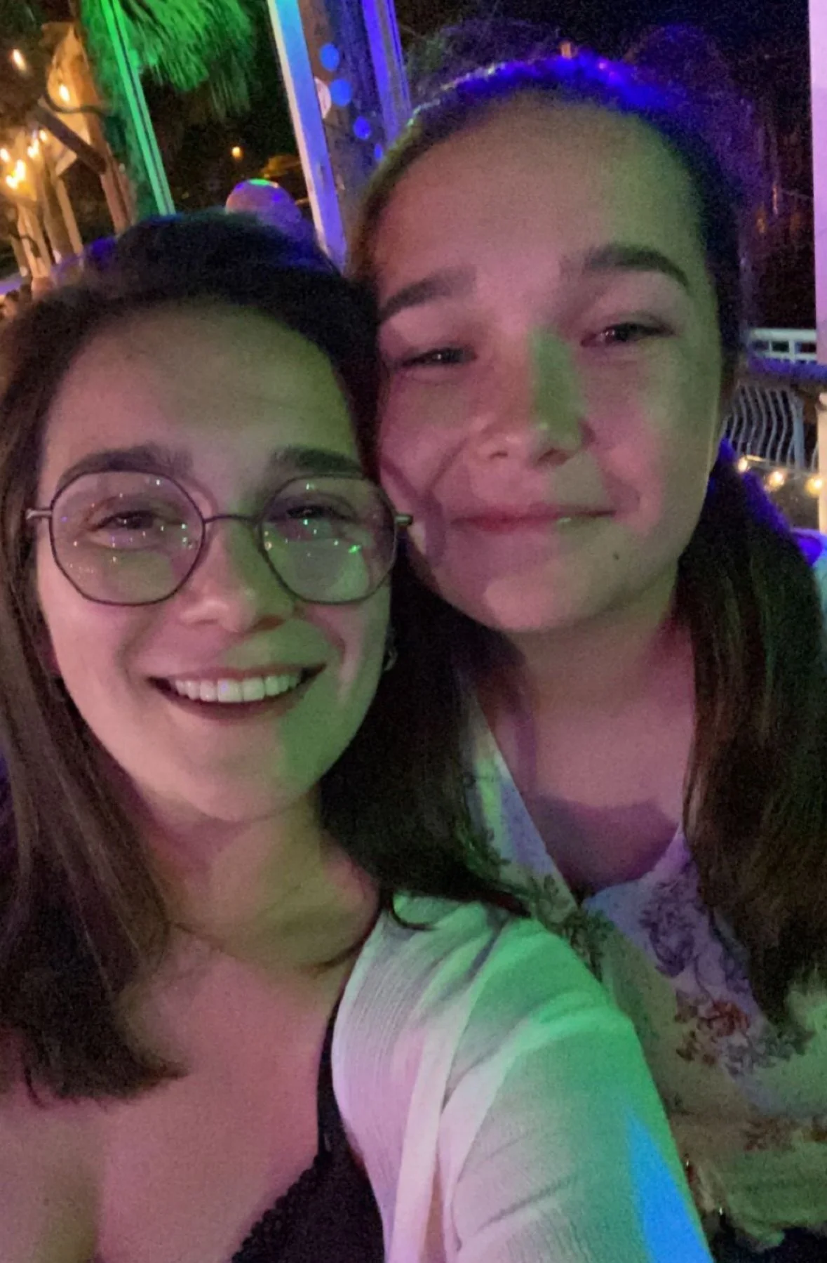 Two women taking a selfie at night, smiling, with colorful lights in the background.