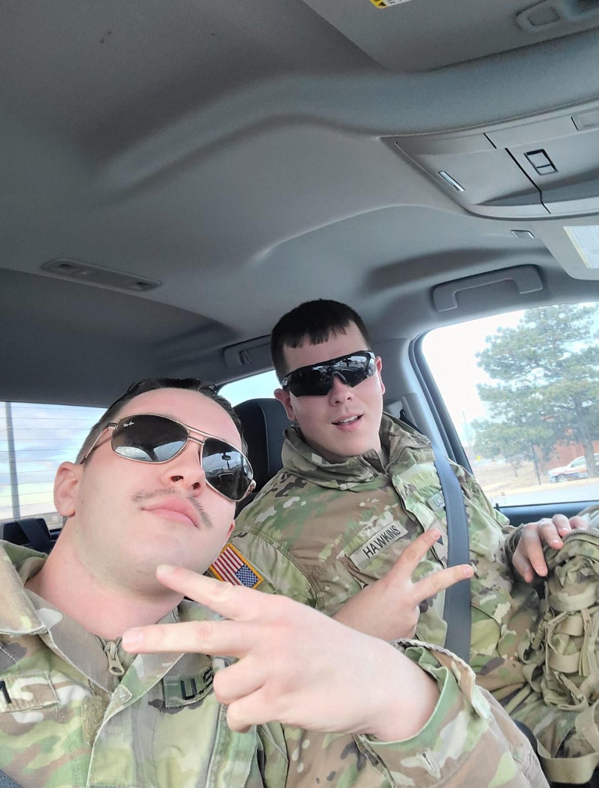 Two soldiers in camouflage uniforms wearing sunglasses, making peace signs inside a vehicle.