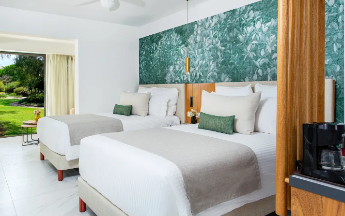 Two double beds in a hotel room with white bedding, green accent pillows, and a teal wallpapered accent wall. A coffee maker is on the right, and a sliding glass door with curtains opens to a garden view.