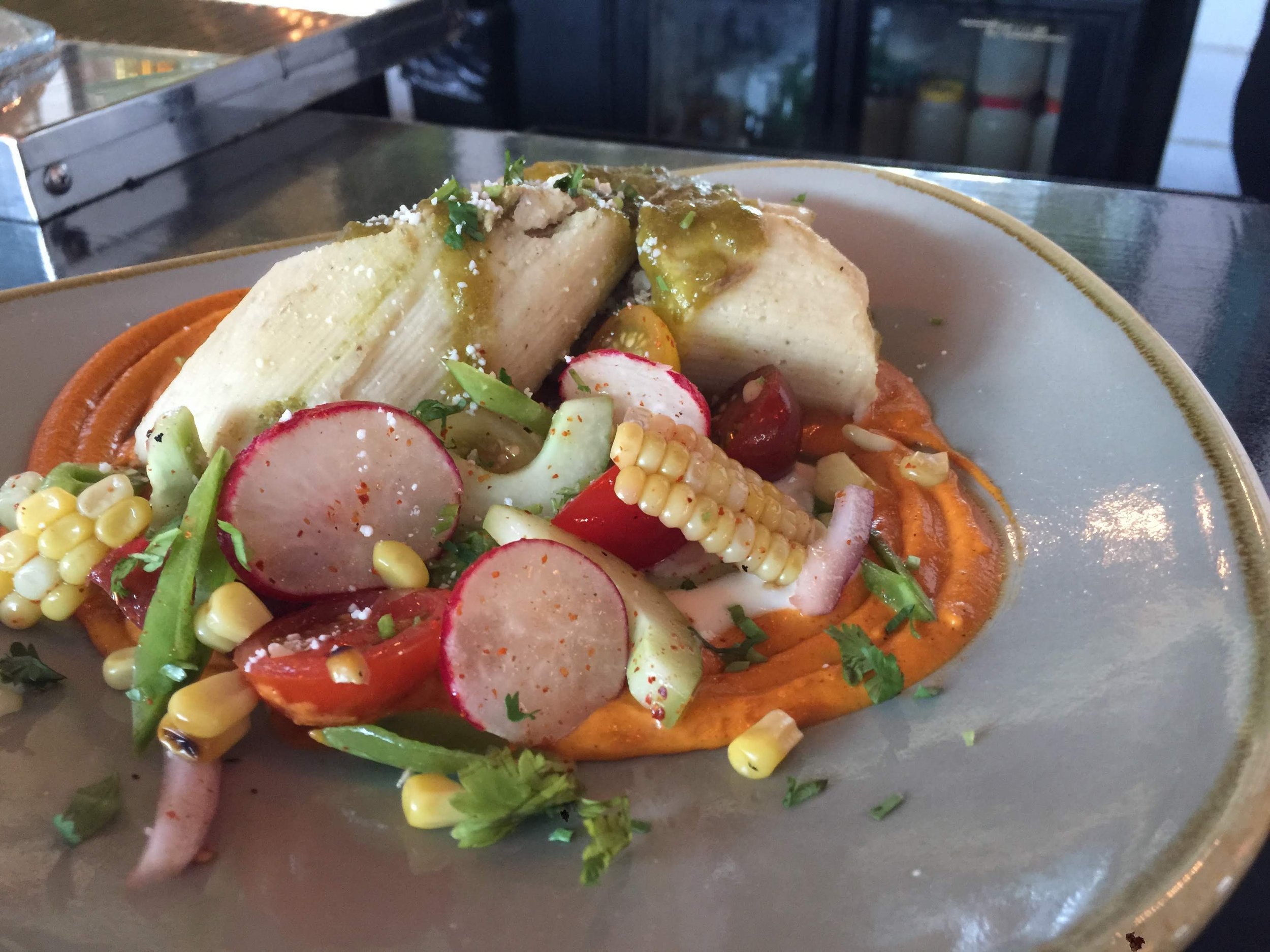 A plate of Mexican food with sliced radishes, corn, cherry tomatoes, and a creamy sauce, served with baked plantains and a dollop of green sauce garnished with chopped herbs.