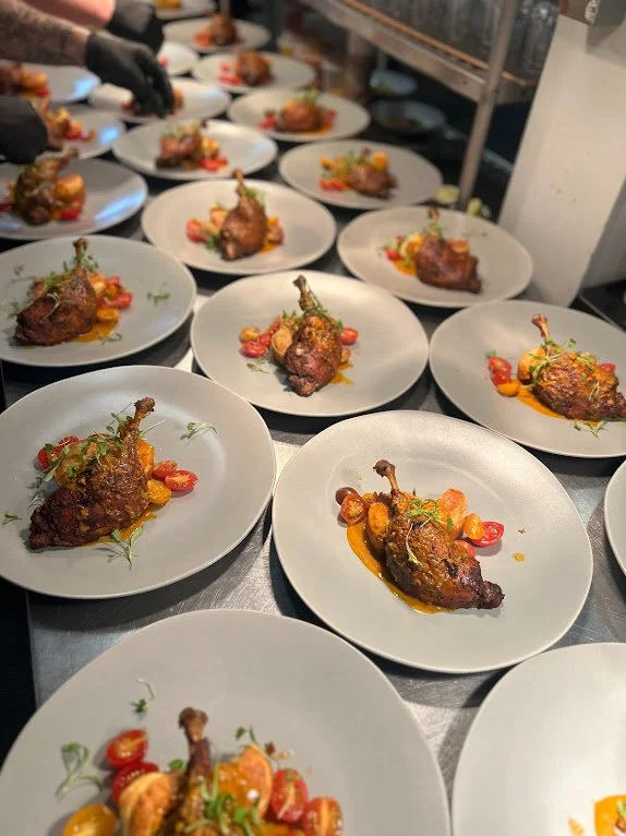 Multiple plated roasted chicken legs garnished with herbs and cherry tomatoes on a kitchen counter.