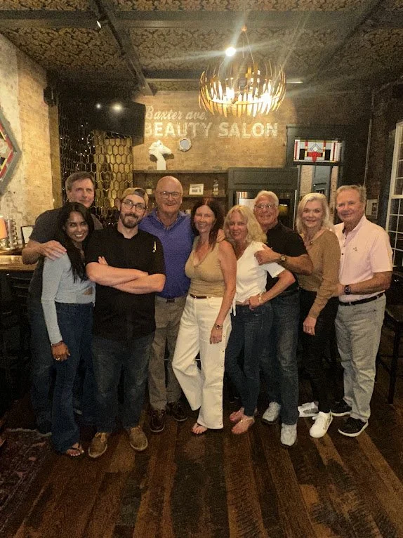 Group of nine people standing together in a cafe or restaurant, smiling at the camera. The background shows a sign that reads 'Beauty Salon' and some decor, with warm lighting and wooden flooring.