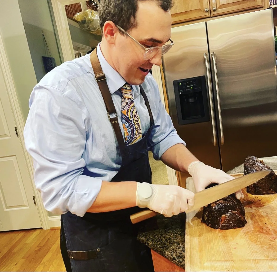 A man wearing glasses, a blue dress shirt, a colorful tie, and a black apron is slicing cooked meat on a wooden cutting board in a kitchen.