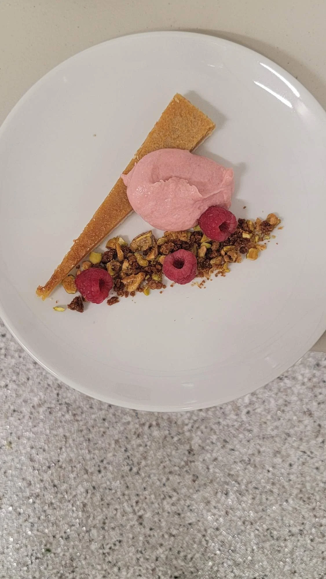 A dessert plate with a slice of tart, pink ice cream, raspberries, and crushed nuts on a white dish.