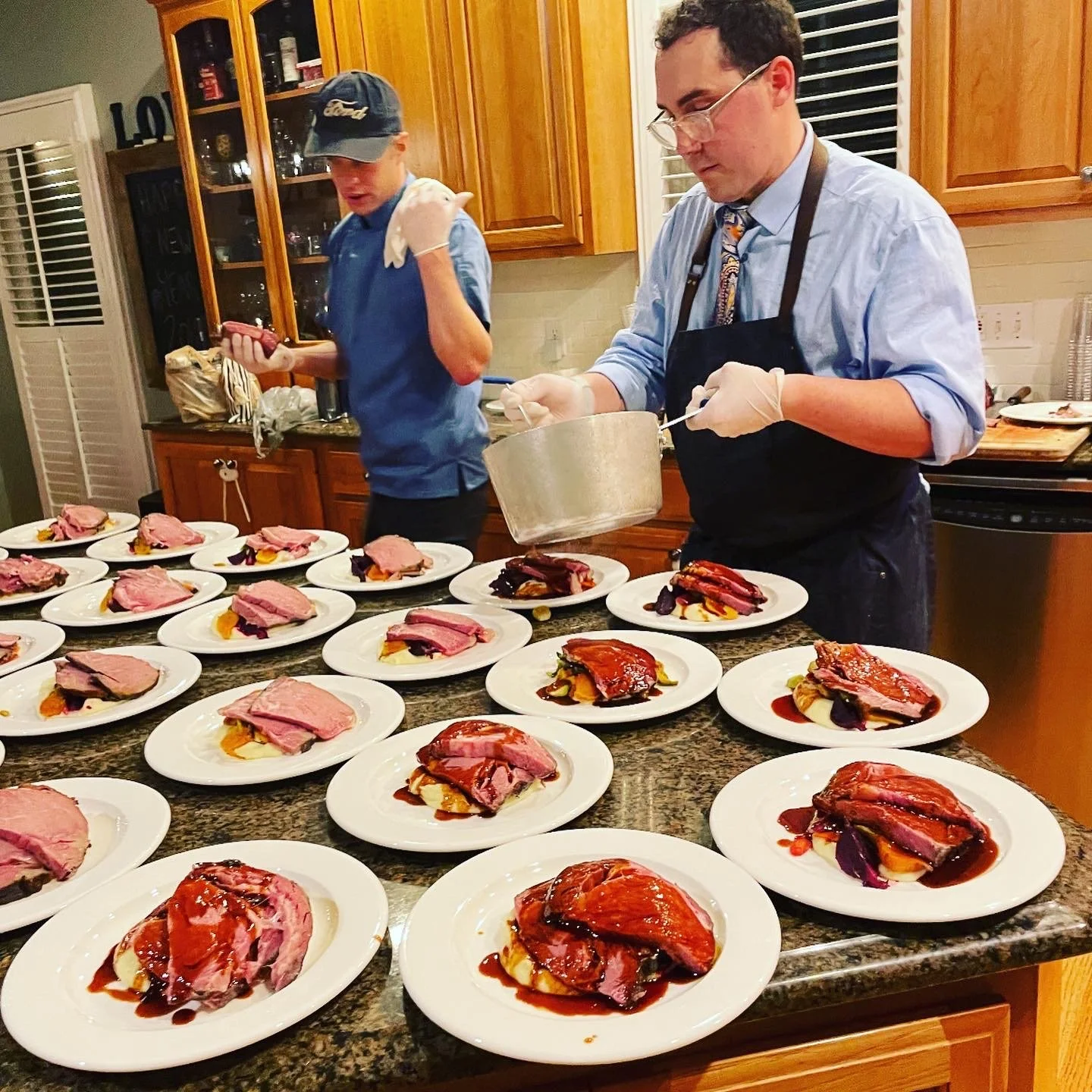 Two men preparing plates of food in a kitchen. One is wearing an apron and handling a pot, while the other, younger man is using a smartphone. Multiple plates with various slices of meat and sauce are arranged on the countertop.