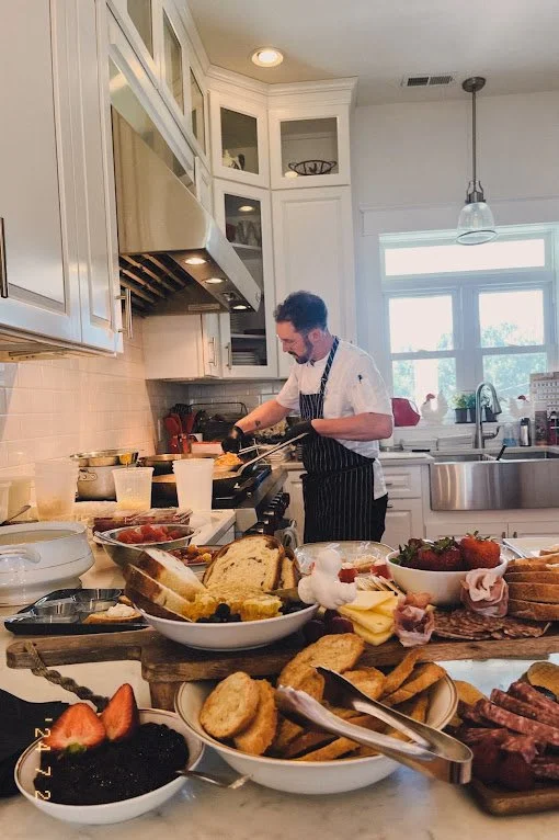 A man preparing food in a kitchen with a breakfast spread in the foreground, including bread, strawberries, blackberries, and various meats.