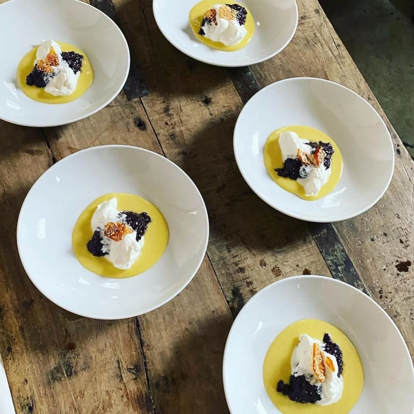 Five white bowls containing a yellow sauce or pudding topped with whipped cream, black caviar, and orange zest, arranged on a rustic wooden table.