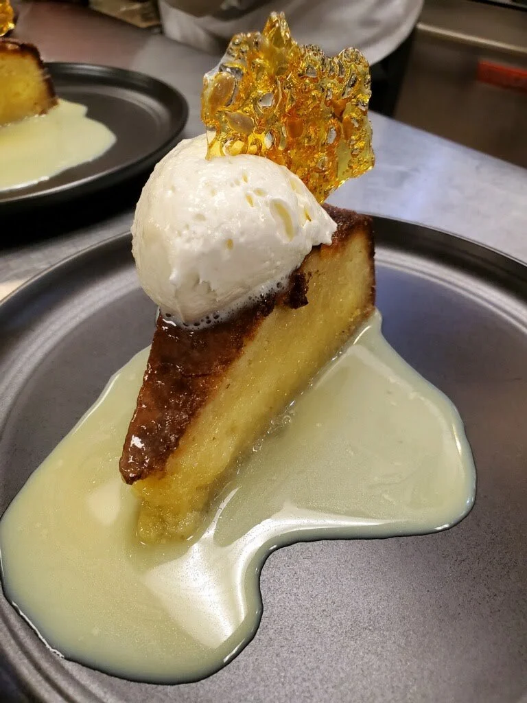 A slice of caramelized cake topped with a scoop of vanilla ice cream, a caramel crisp, and drizzled with a light-colored sauce, served on a black plate.