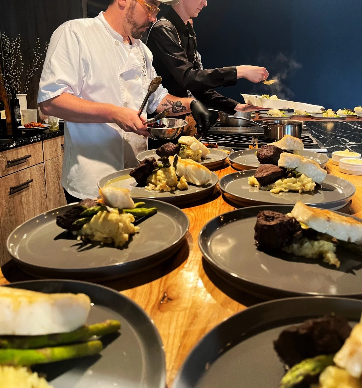 Chefs preparing plated gourmet dishes with steak, mashed potatoes, asparagus, and other vegetables in a professional kitchen.