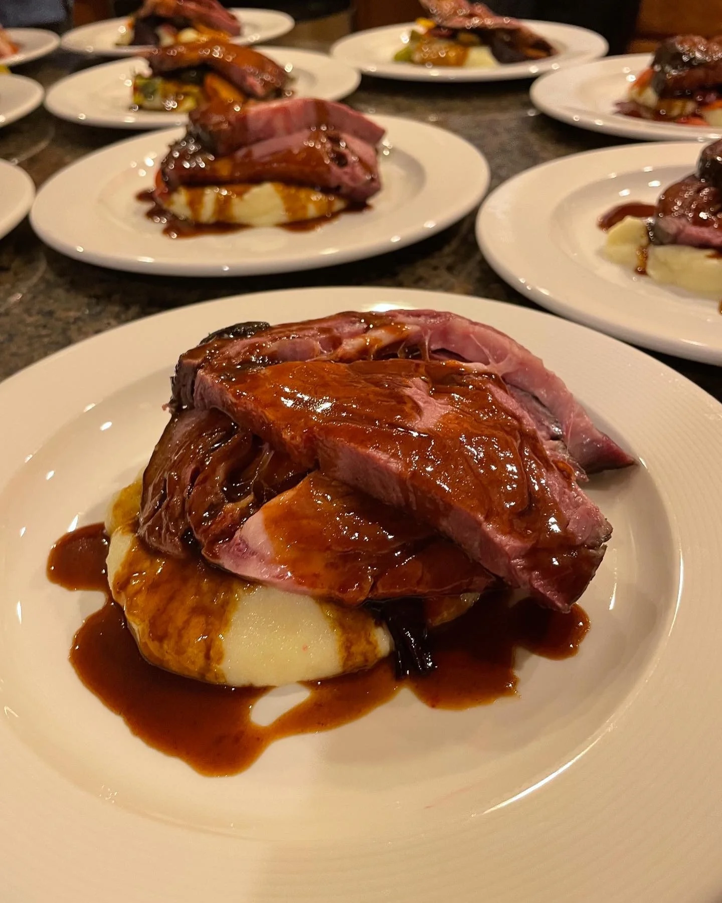 Multiple white plates with servings of roast beef, mashed potatoes, and vegetables, topped with brown gravy, arranged on a dark countertop.