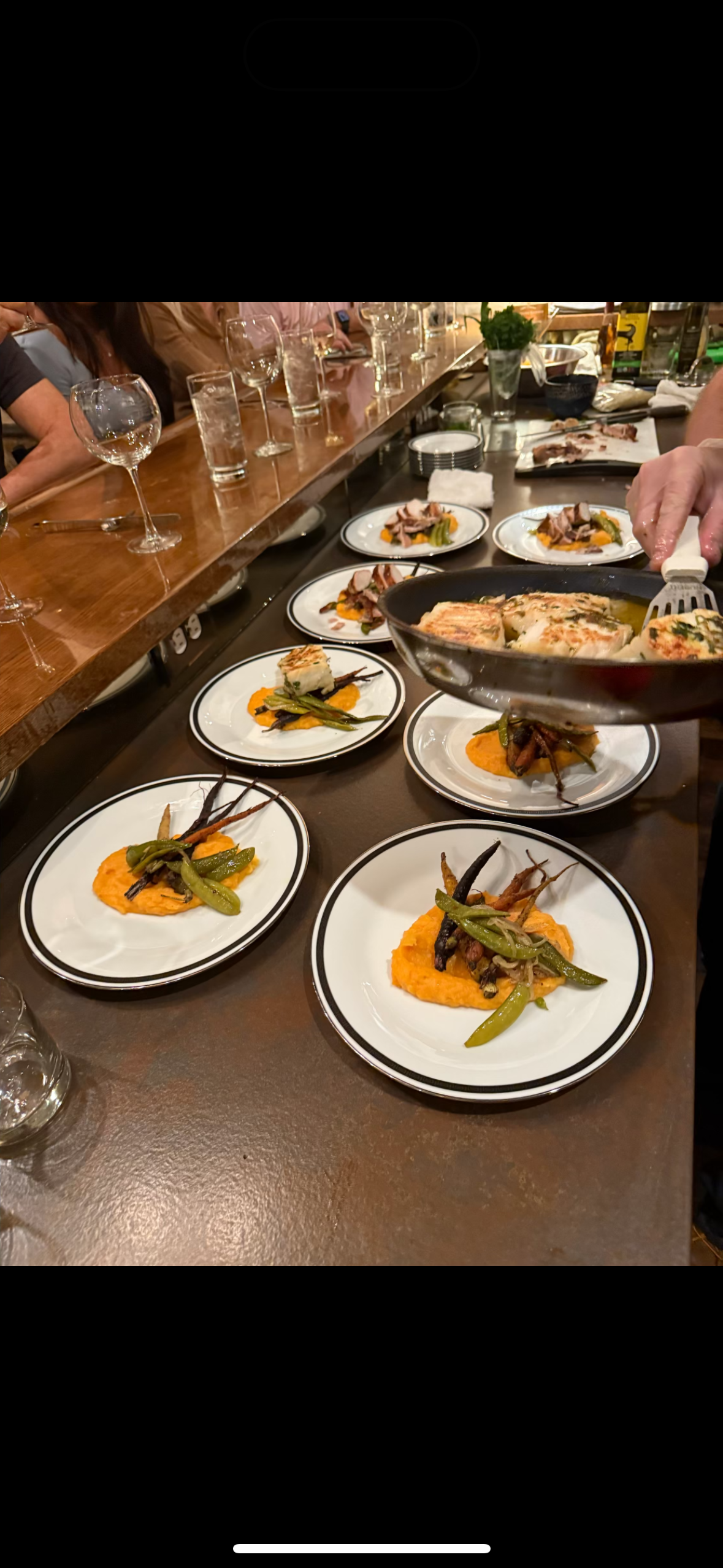 Plates of food with roasted carrots and green beans, and a piece of fish or meat, on a dark table. In the background, a person is holding a skillet with cooked food, and a line of empty wine glasses and water glasses is on a wooden bar counter.