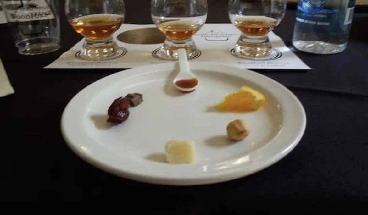A white plate with small samples of food and a small spoon with sauce, placed on a black table. There are three glasses of amber-colored liquid and two bottles of water in the background.
