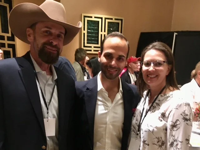 Three people smiling at an indoor event, with a man in a cowboy hat on the left, a man in the middle, and a woman on the right, surrounded by other attendees.