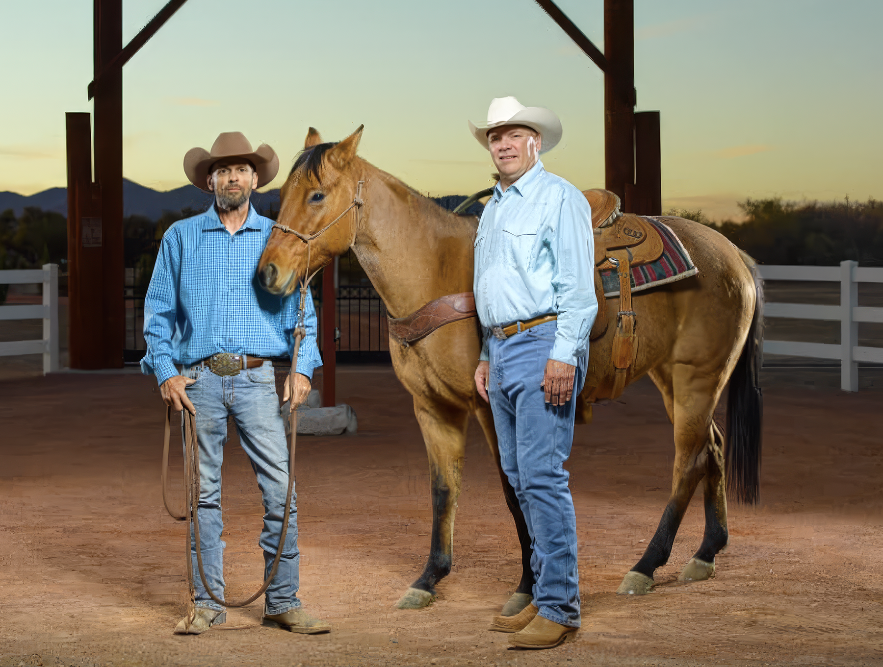 Two men in cowboy attire, standing beside a brown horse with a saddle, inside a covered riding arena at sunset.