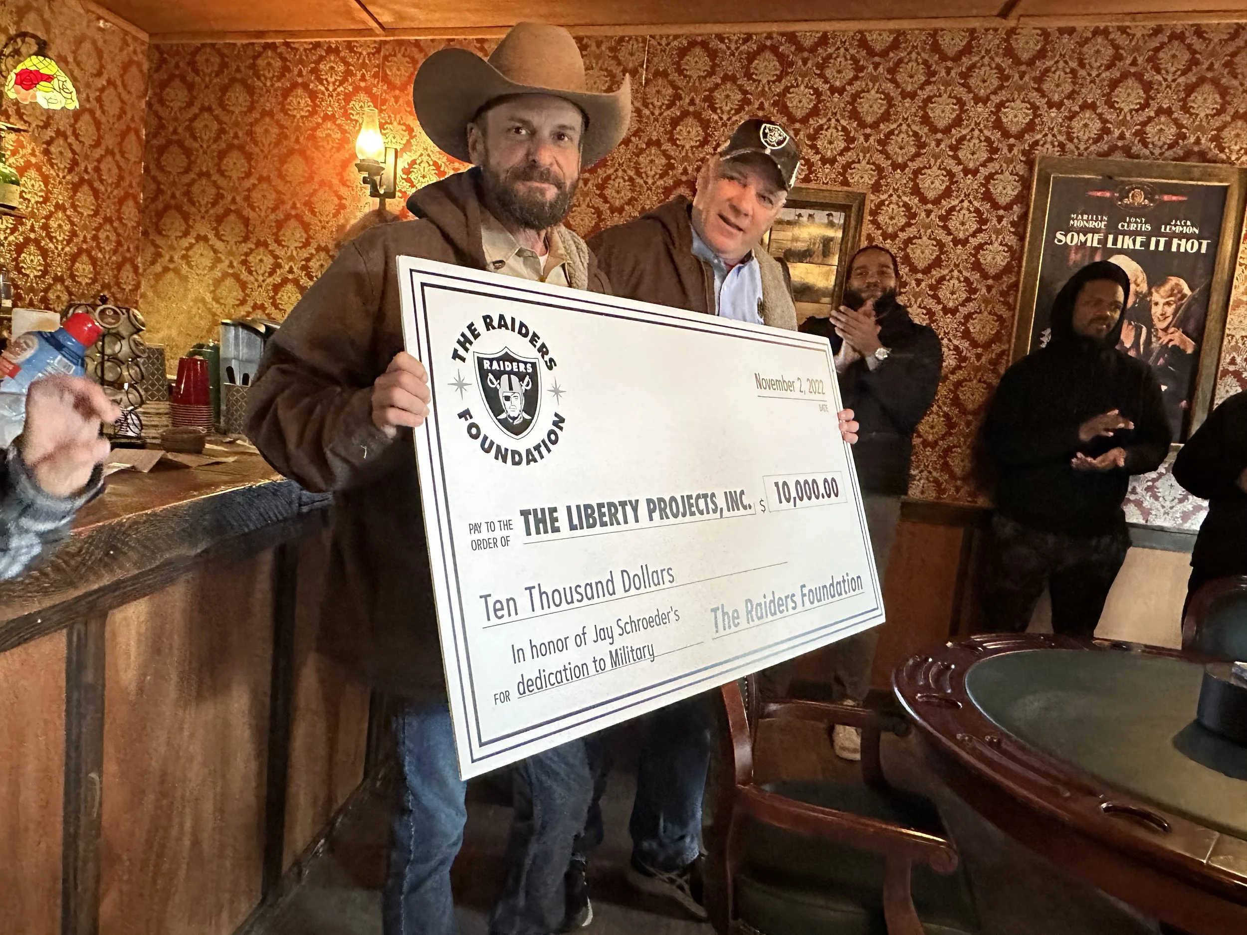 A man dressed in a cowboy hat and brown jacket holds a large check for $10,000 made out to The Liberty Projects, Inc. Standing beside him is another man wearing a cap and a jacket. Behind them, several people are clapping in a warmly decorated room with patterned wallpaper, artwork on the wall, and a poker table in the foreground.