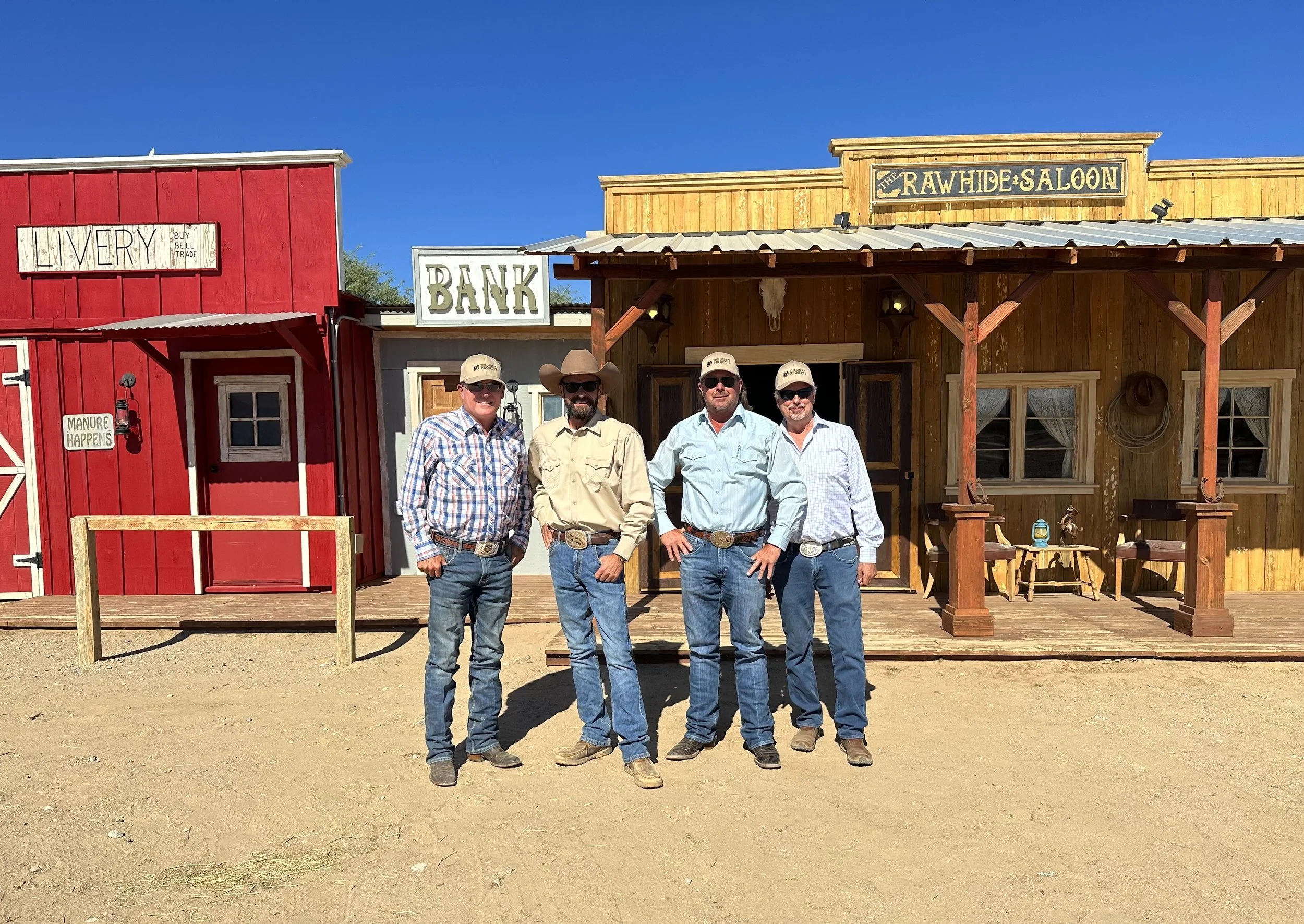 Four men dressed in cowboy attire, standing in front of a rustic Western-themed building with signs reading 'The Rawhide Saloon,' 'Bank,' and 'Livery.' The background is clear blue sky.