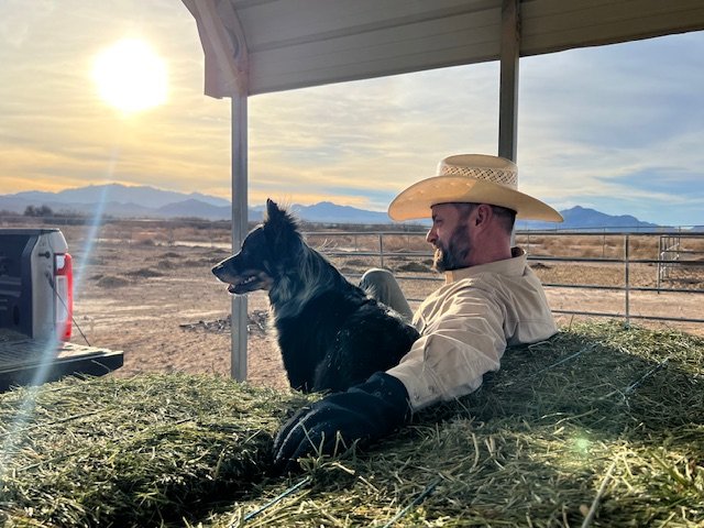 Man wearing cowboy hat and beige shirt relaxing on a farm with dog, open field, mountains in the background, and sunset sky.