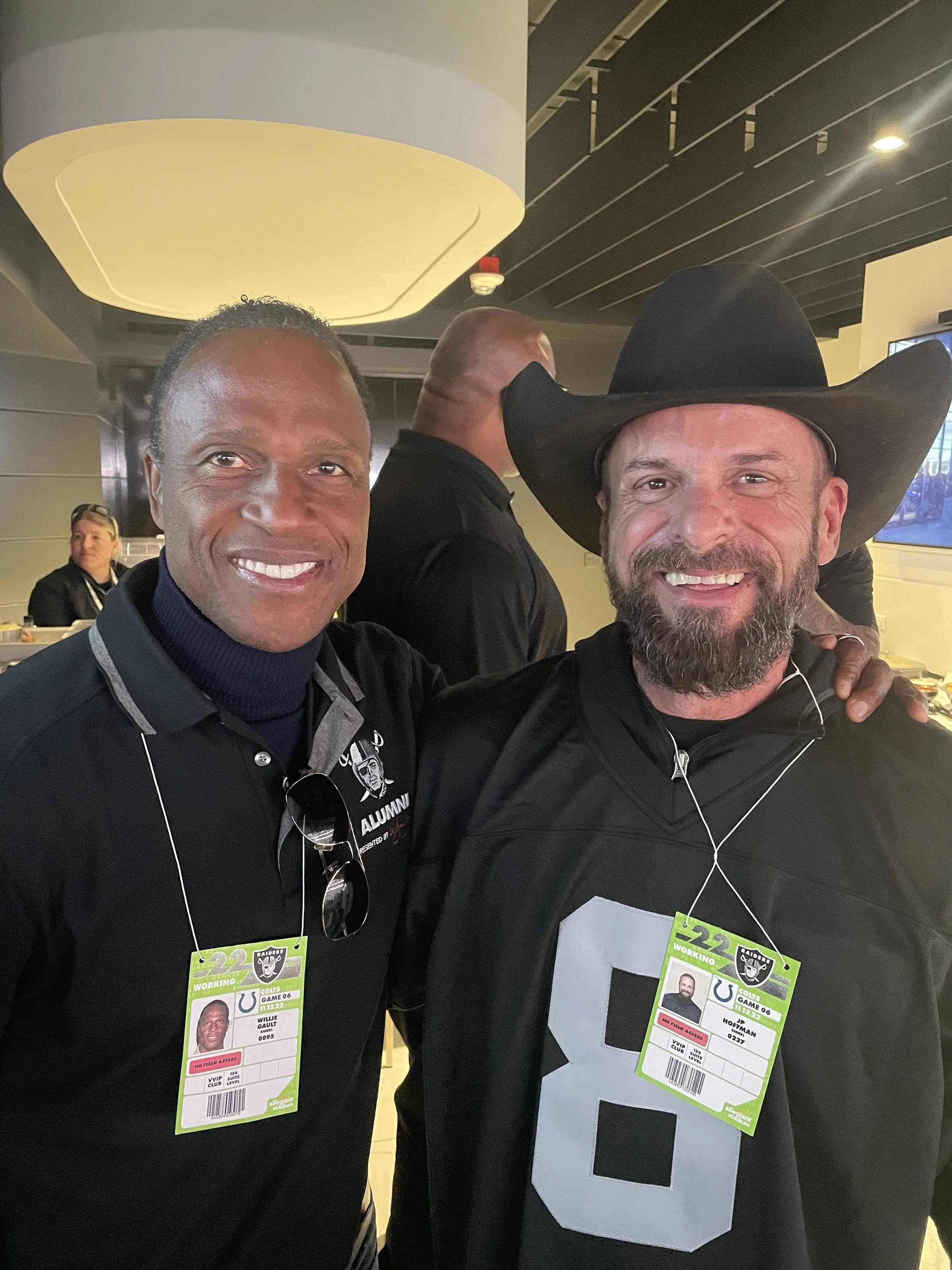 Two men smiling at a sports event, one wearing an Oakland Raiders jacket and a cowboy hat, and the other wearing a Raiders shirt and a badge. They are in an indoor stadium or arena.
