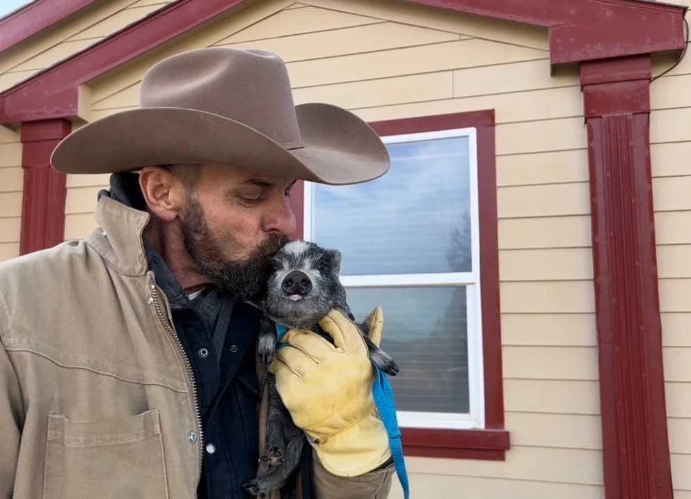 J.P. Hoffman in a cowboy hat and tan jacket holding a small black and white piglet outside a yellow house with red trim.