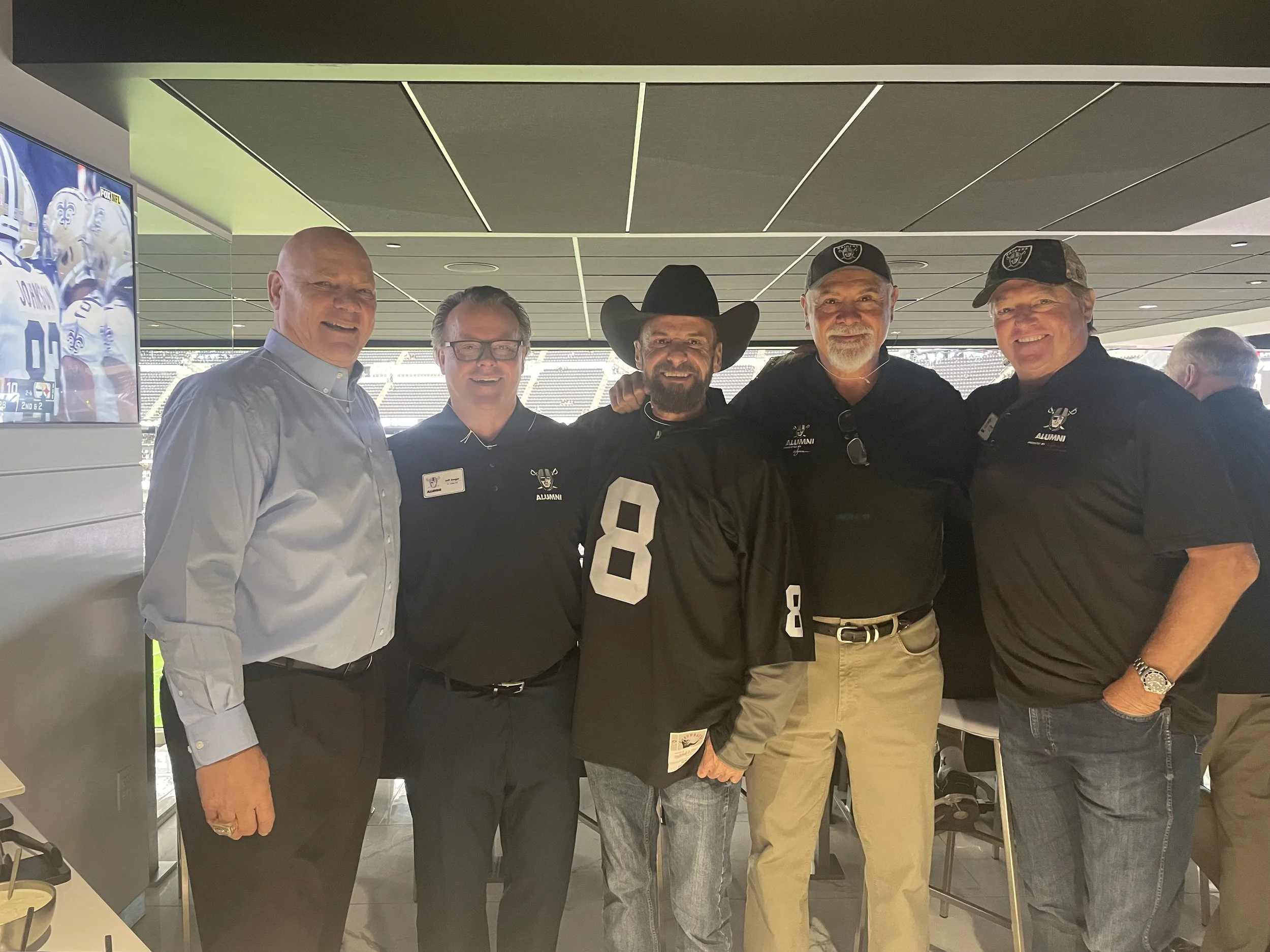 Group of five men standing in a stadium, some wearing Oakland Raiders caps and shirts, smiling at the camera.