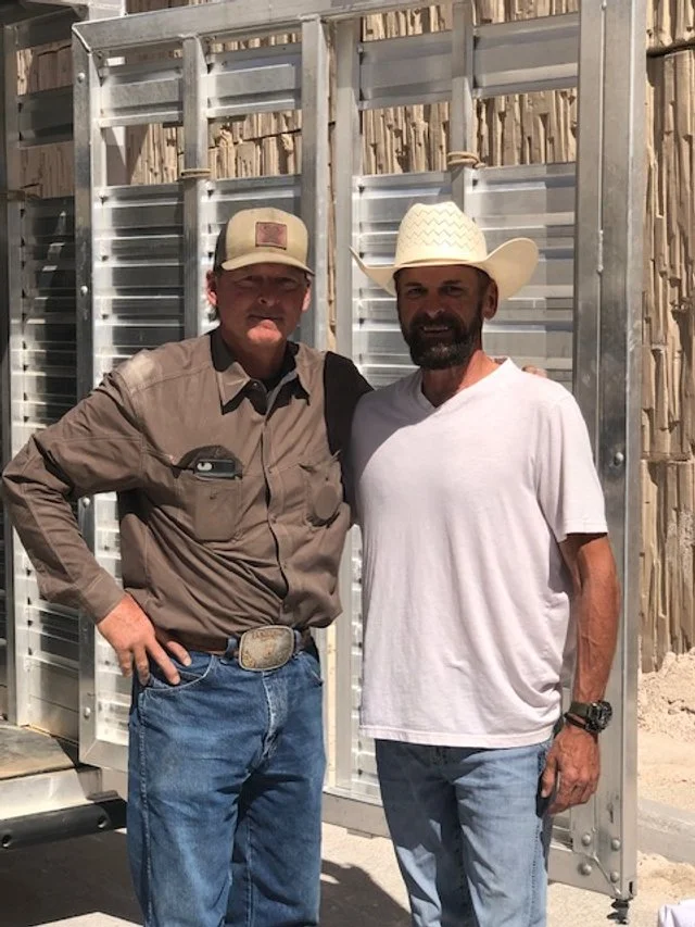 Two men standing outdoors in front of a metal fence with wooden slats. One man wears a cowboy hat, a white t-shirt, jeans, and a watch, smiling at the camera. The other man wears a cap, a button-up shirt, jeans, and has a hand on his hip, also smiling.