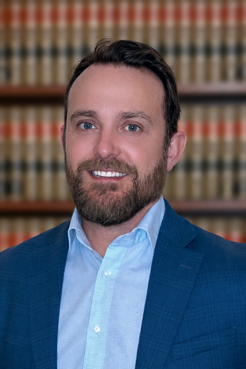 A man with brown hair and a beard, wearing a blue blazer and light blue shirt, smiling in front of a bookshelf filled with legal books.