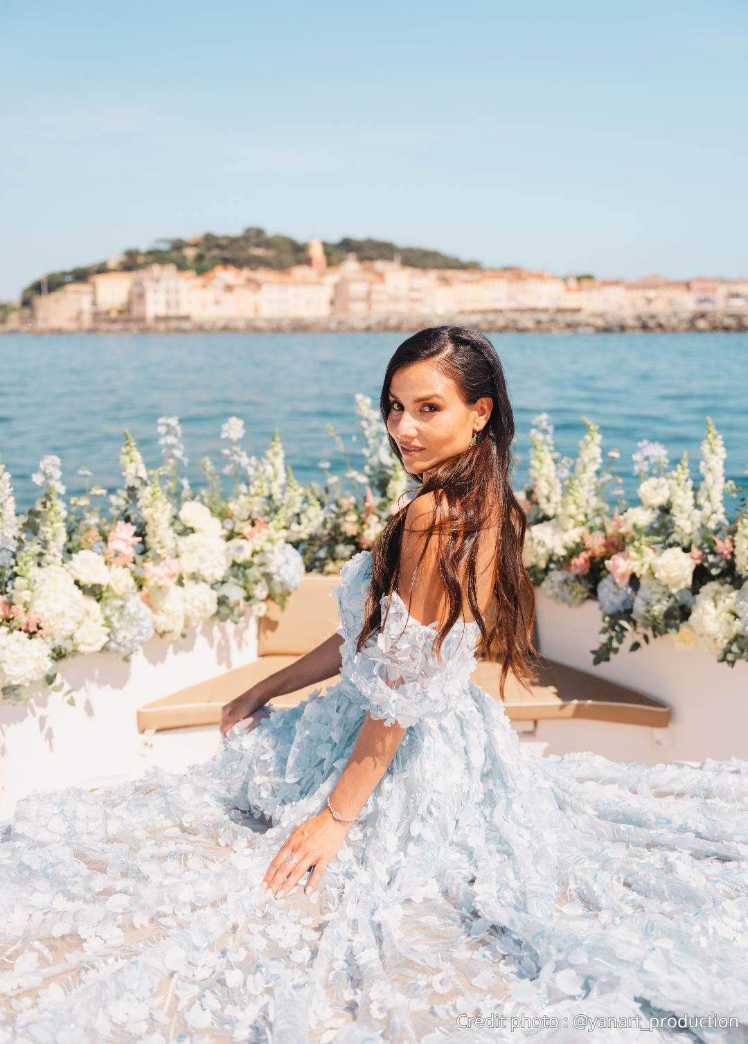 Une femme en robe de mariage blanche assise dans un décor floral près de l'eau avec une ville en arrière-plan.