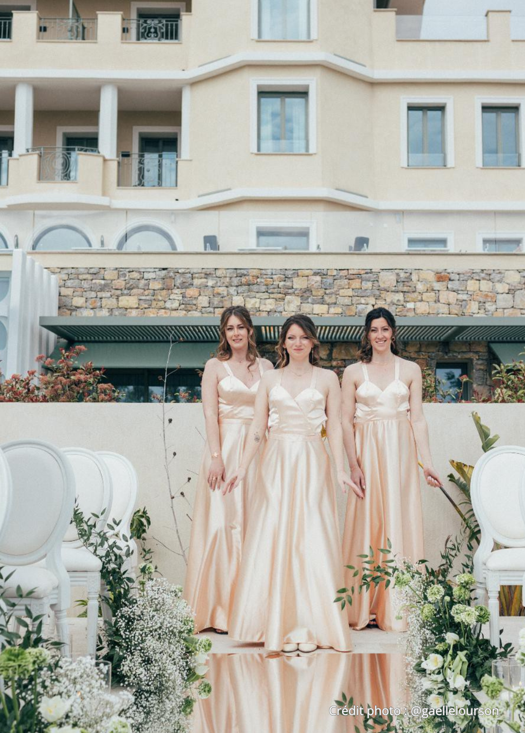 Trois femmes portant des robes de mariage roses pâles, debout dans un décor de mariage avec des fleurs et des chaises blanches, devant un bâtiment moderne beige et pierre.