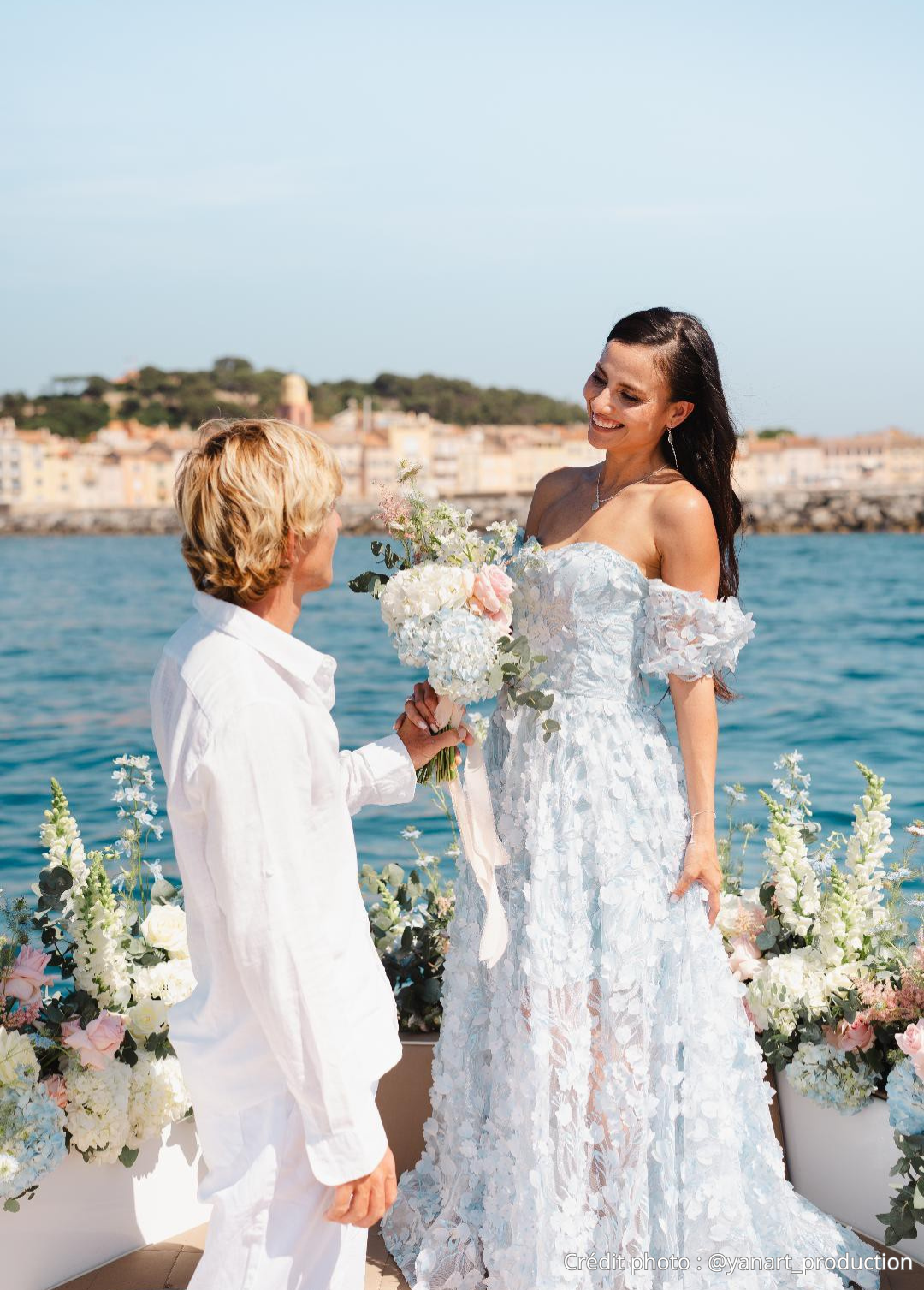 Une femme en robe de mariée tenant un bouquet de fleurs, souriante, lors d'une cérémonie en extérieur près de l'eau, avec un homme lui faisant une demande en mariage.