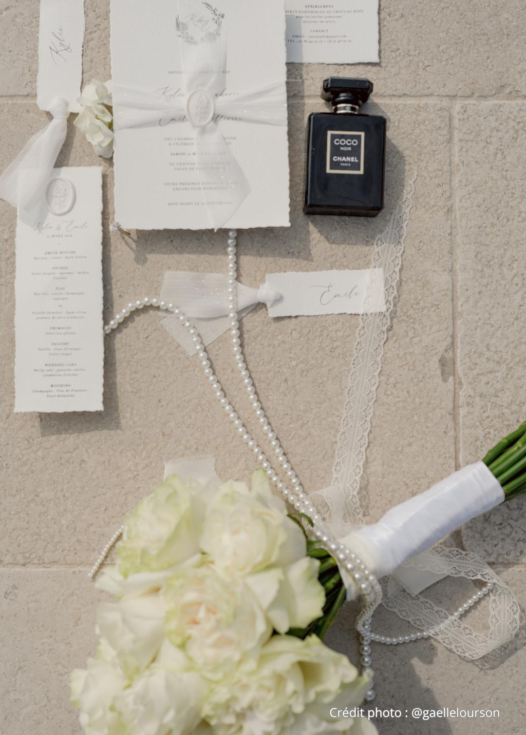 Accessoires de mariée blancs : robe, bouquet, perles et faire-part posés sur un mur en pierre.