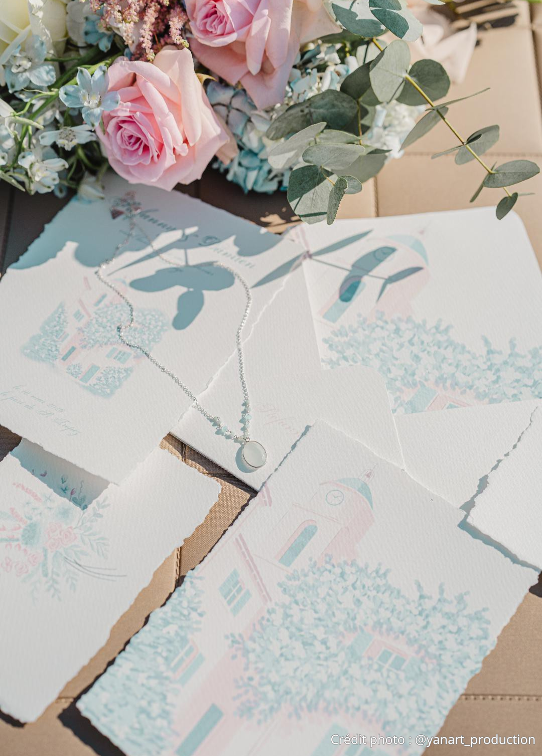 Une composition florale avec des roses roses, des fleurs blanches, et des feuillages, accompagnée de papiers décoratifs et d'un collier avec un pendentif, sur une surface en cuir.