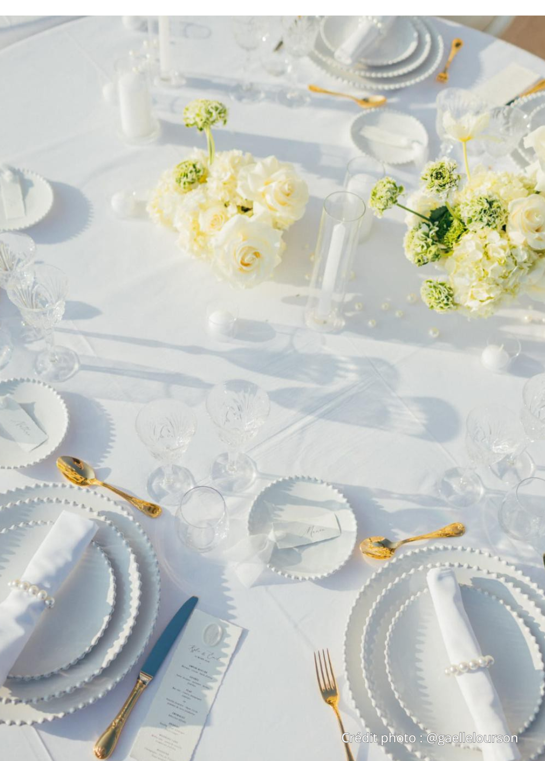 Table de mariage élégante décorée de fleurs blanches, vaisselle blanche, assiettes avec perles, couverts dorés, verres en cristal, et une carte de menu. La table est en plein soleil.