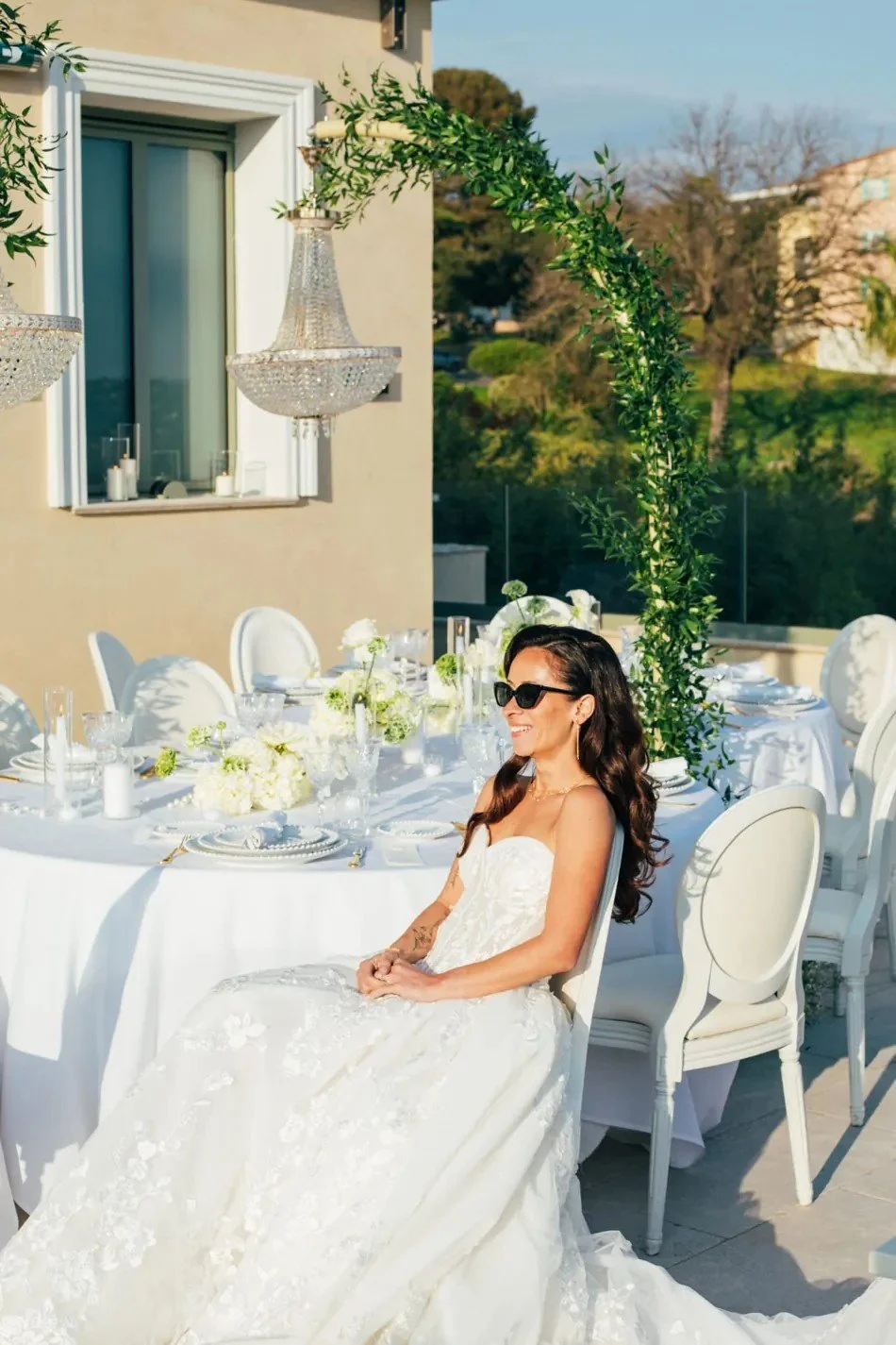 Mariée assise à une table blanche décorée, lustre en cristal et arche de verdure.