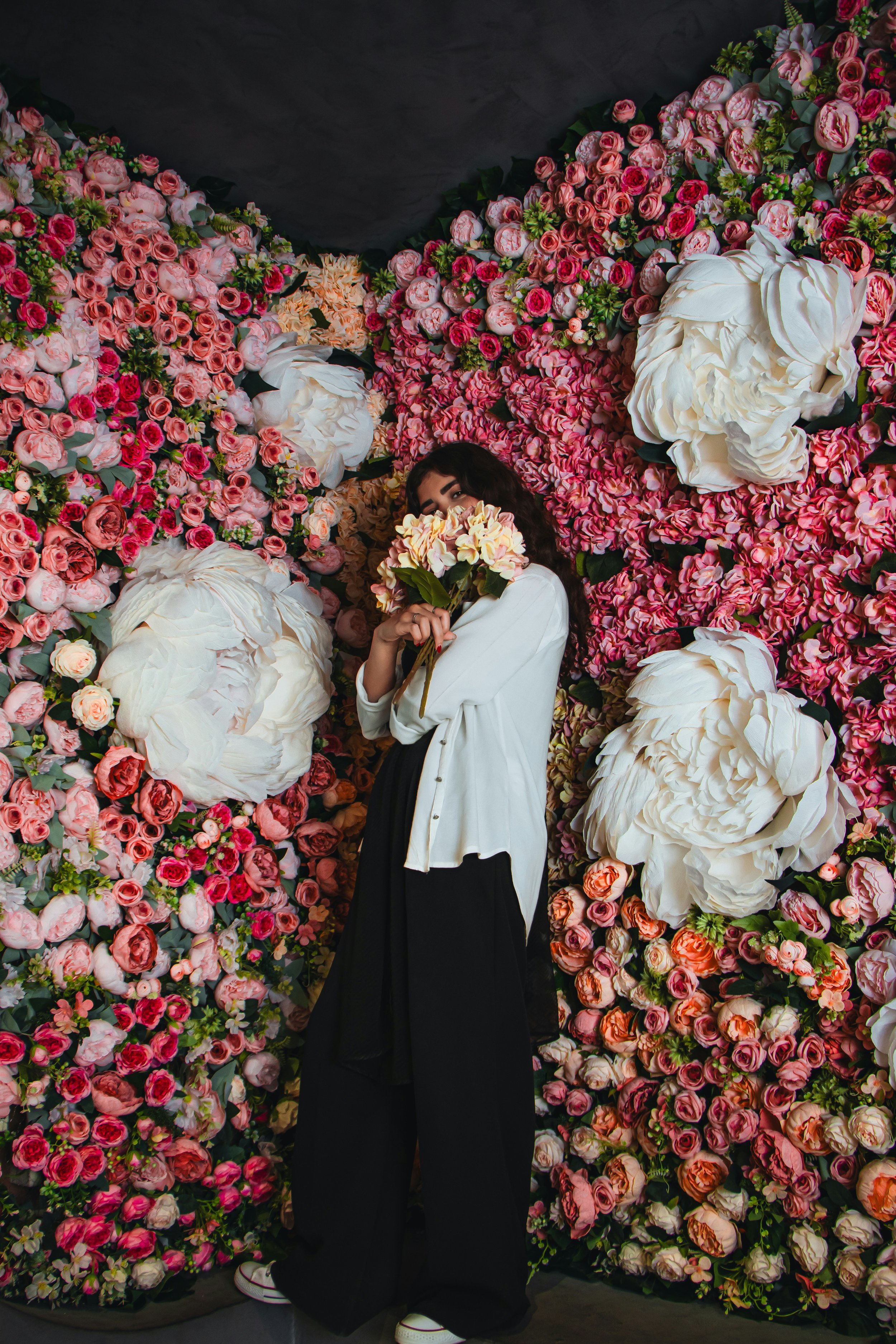 Une femme tenant un bouquet de fleurs, entourée d'un mur de fleurs roses, blancs et orange.
