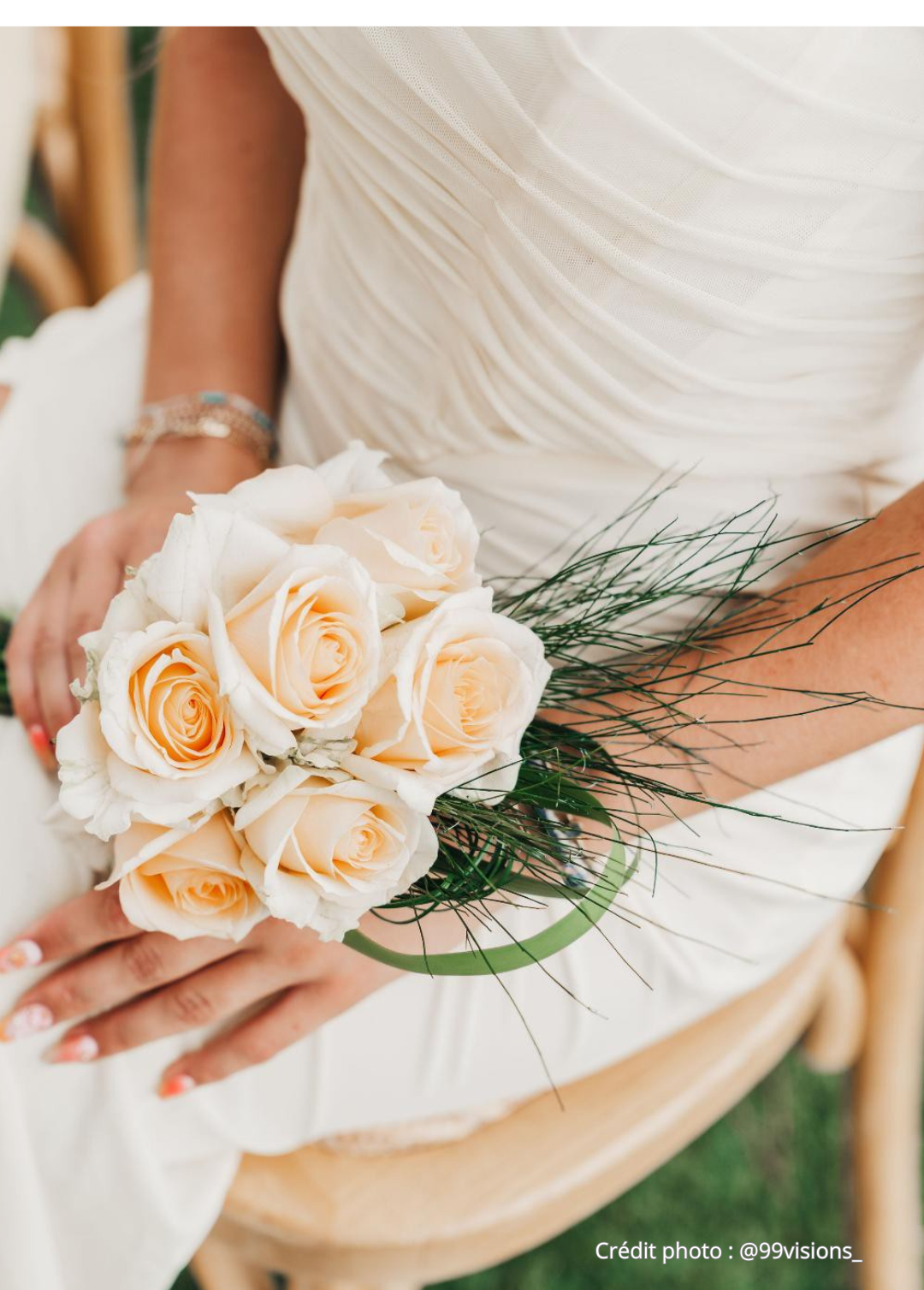 Une personne en robe blanche de mariage tient un bouquet de roses crème avec des feuillages verts.