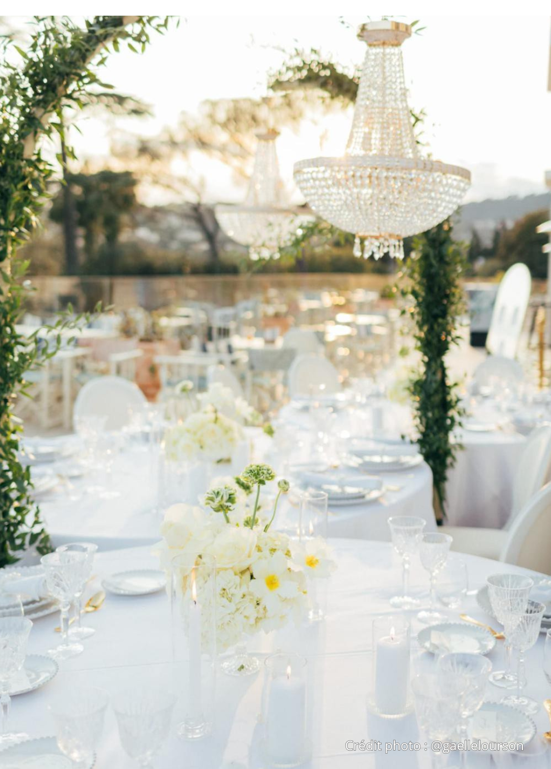 Table décorée pour un événement en plein air, avec chandeliers en cristal, fleurs blanches et vaisselle élégante sous un ciel ensoleillé.