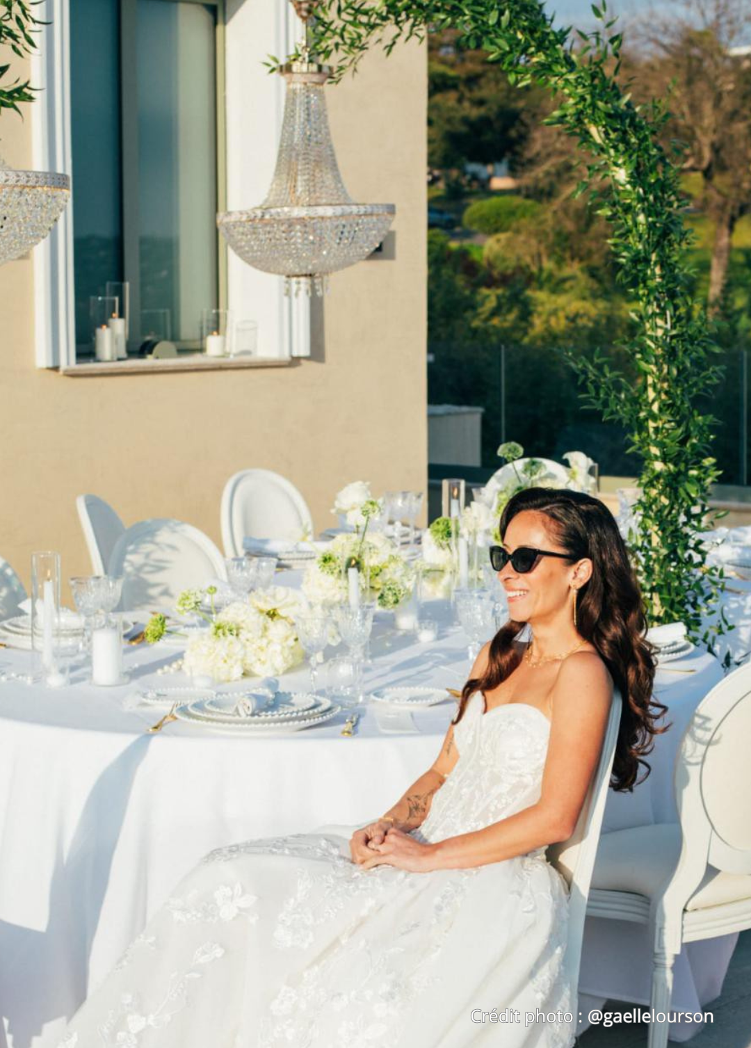 Une femme en robe de mariée assise à une table décorée de fleurs blanches lors d'un événement en plein air.