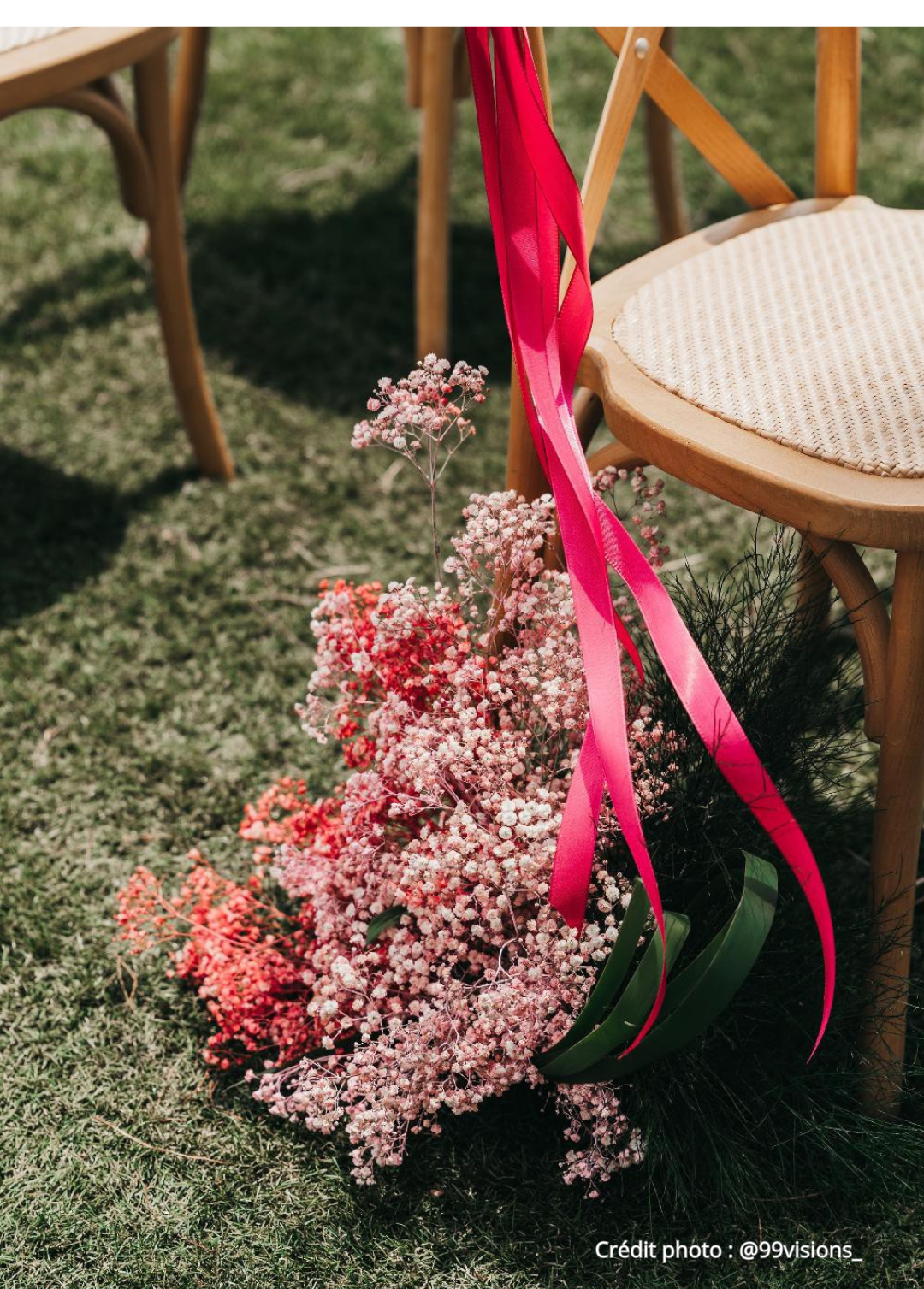 Une chaise en bois avec un siège en paille, un bouquet de fleurs roses et rouges avec un ruban rose, posé sur l'herbe lors d'un événement en plein air.