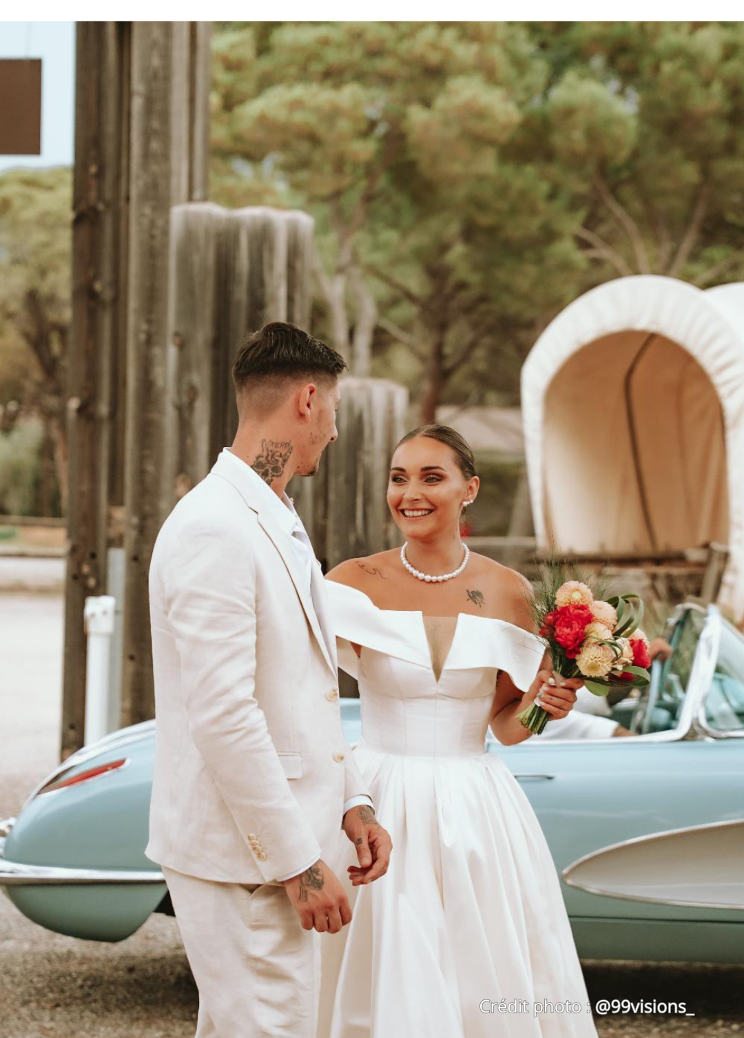 Un couple de mariés souriants, la femme tenant un bouquet de fleurs, dans une ambiance de mariage en plein air, avec une calèche en arrière-plan.