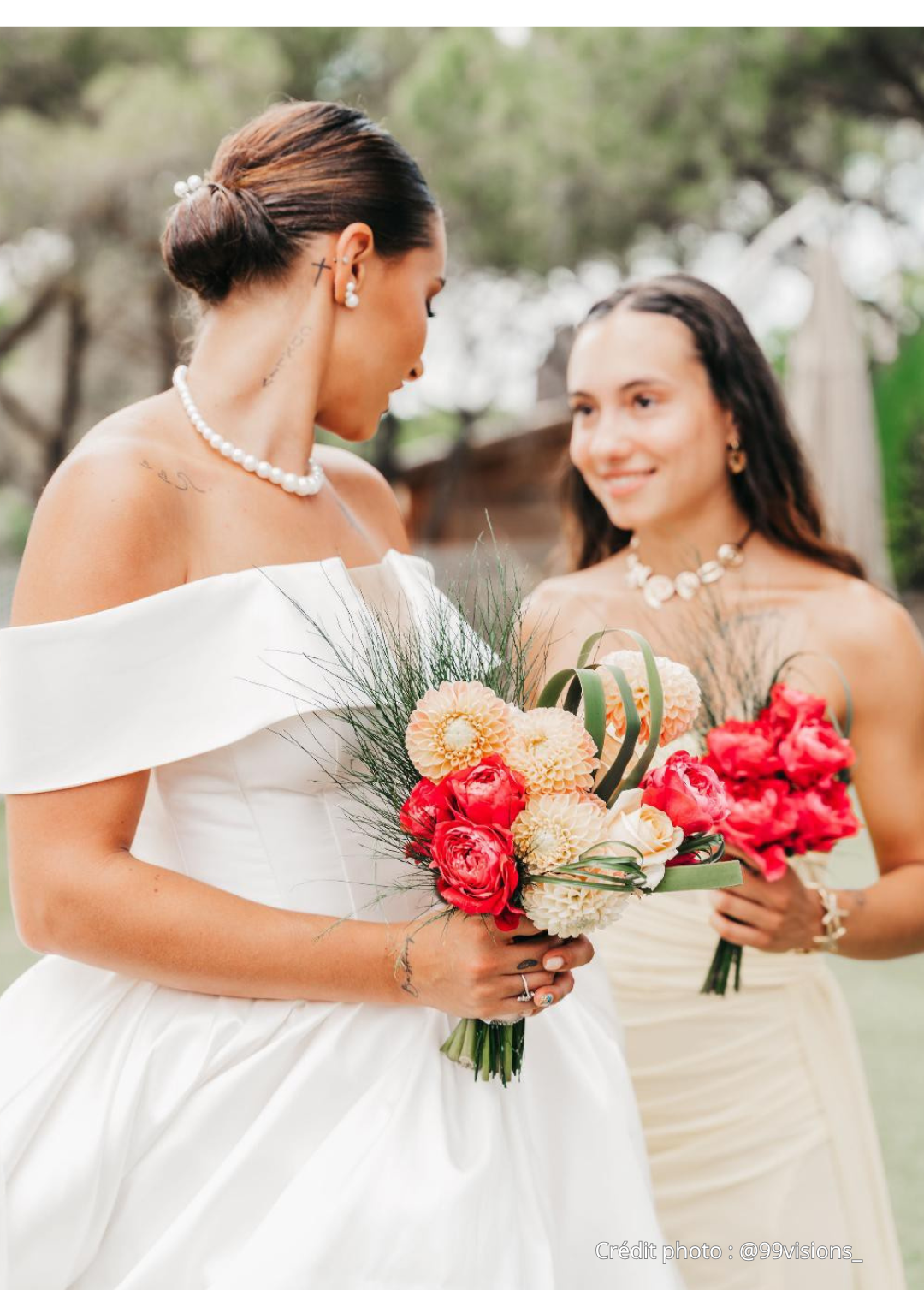 Deux femmes lors d'un mariage, l'une en robe de mariée blanche tenant un bouquet de fleurs roses, l'autre en robe beige avec un collier de coquillages, dans un cadre extérieur avec des arbres en arrière-plan.