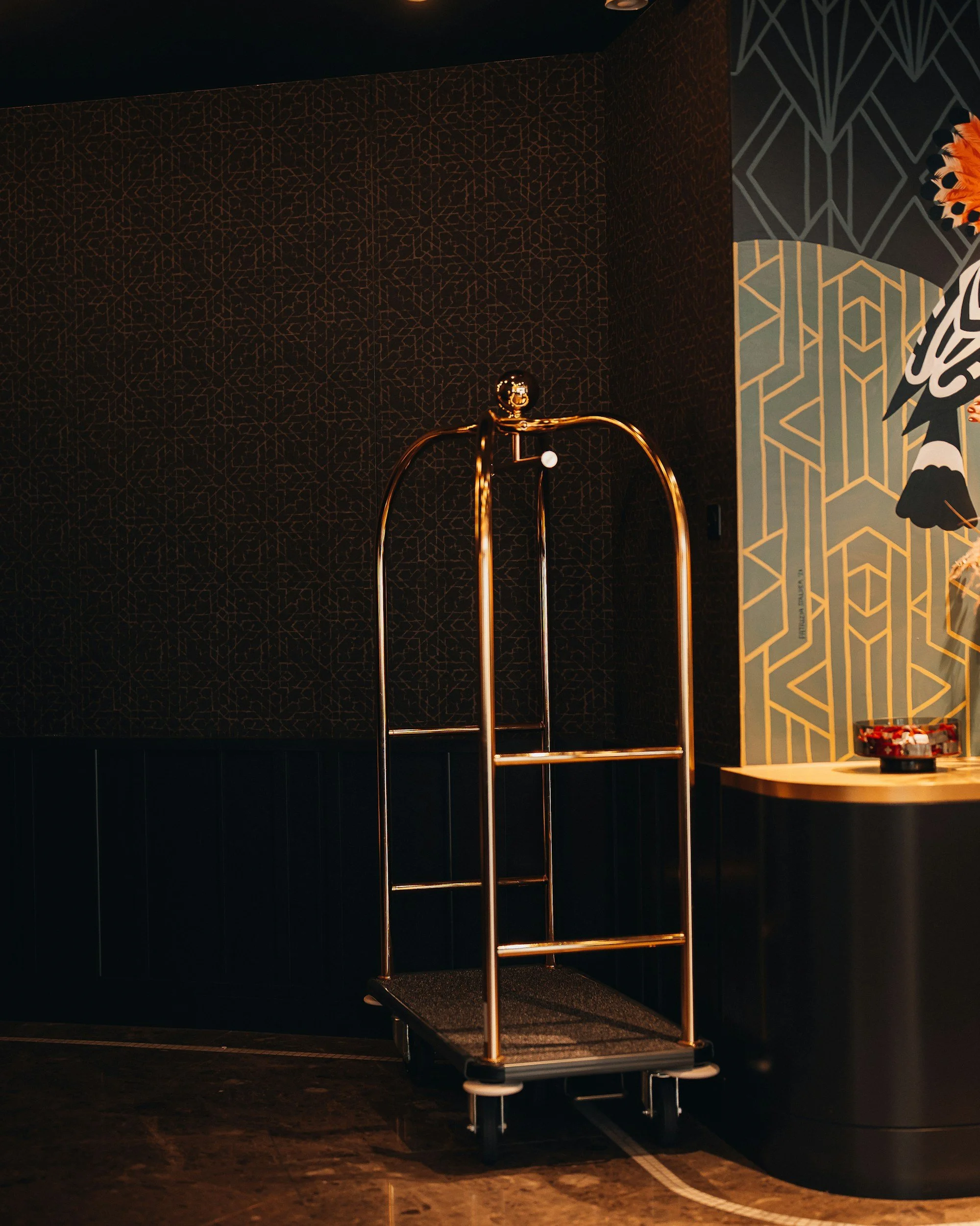 A hotel luggage cart made of metallic gold with a step platform at the bottom, situated against a dark-colored wall with geometric patterns, partially visible artwork, and a side table with small items on it.