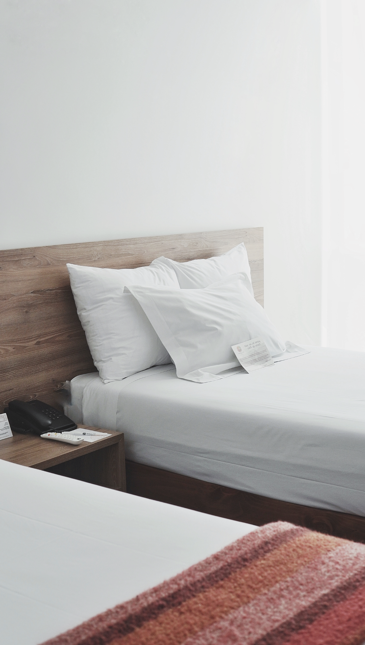 A hotel room with a bed, two pillows, a remote control, a telephone, and some papers on a wooden bedside table, with white walls and minimal decor.