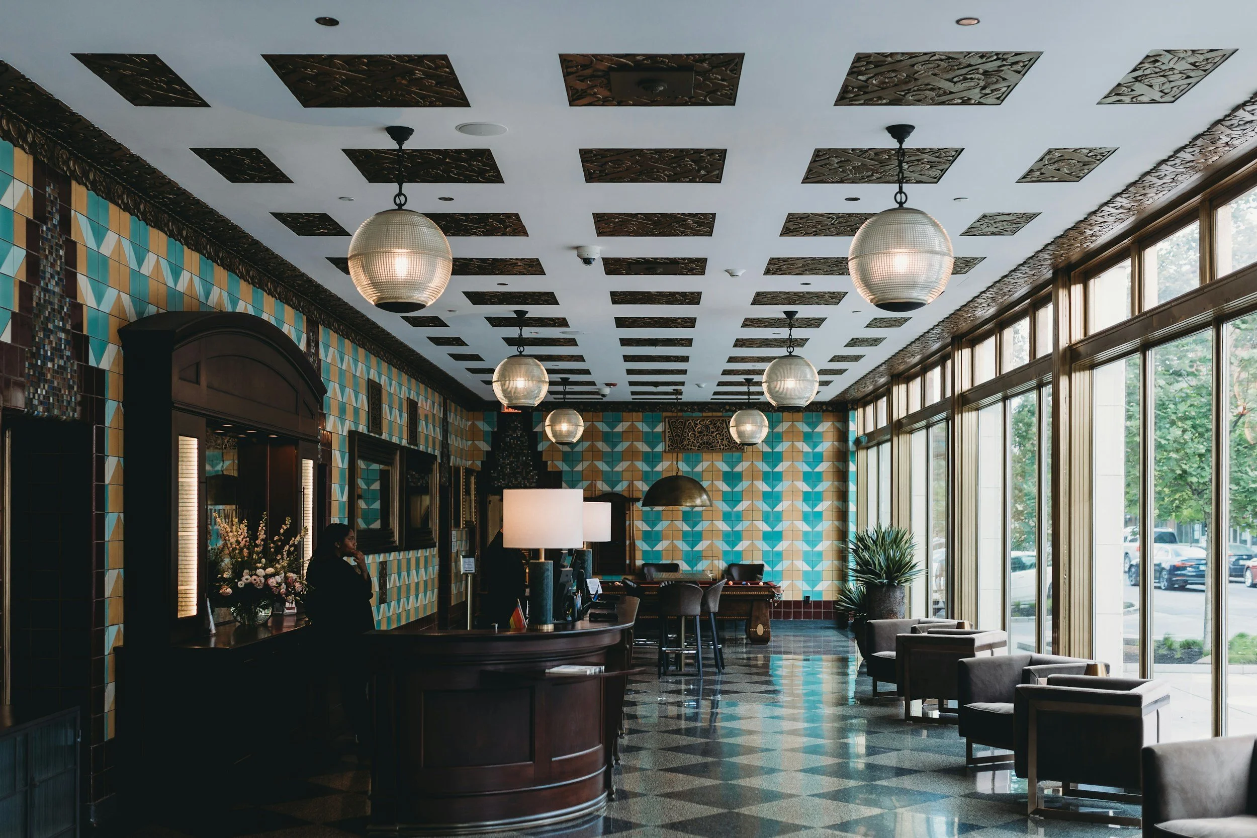 Hotel lobby with geometric wall tiles, large windows, seating, reception desk, and pendant lights.
