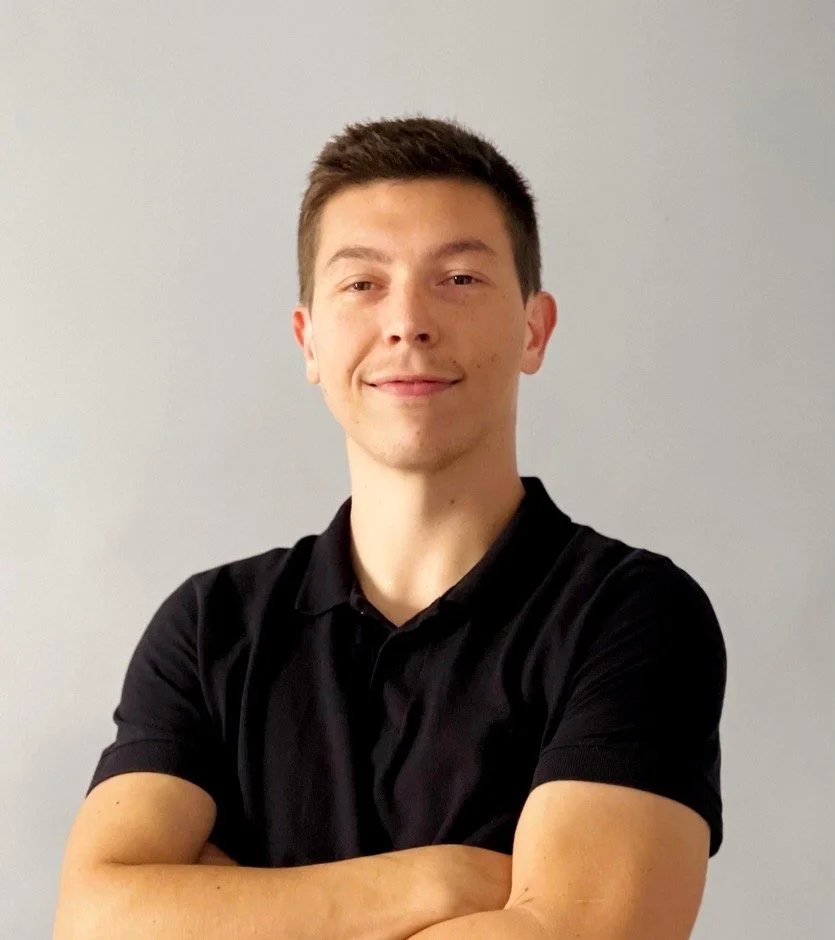 A young man with short brown hair, wearing a black polo shirt, standing with arms crossed against a plain light gray background.