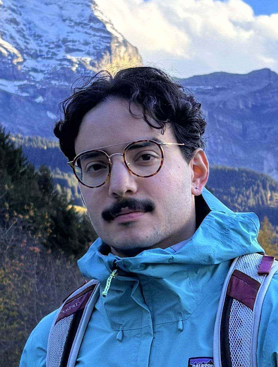 A man with glasses and a mustache outdoors with mountains and trees in the background.