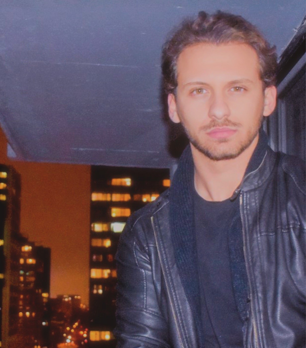 A young man with short, wavy brown hair and a beard posing on a balcony at night, with a cityscape and illuminated windows in the background, wearing a black leather jacket.