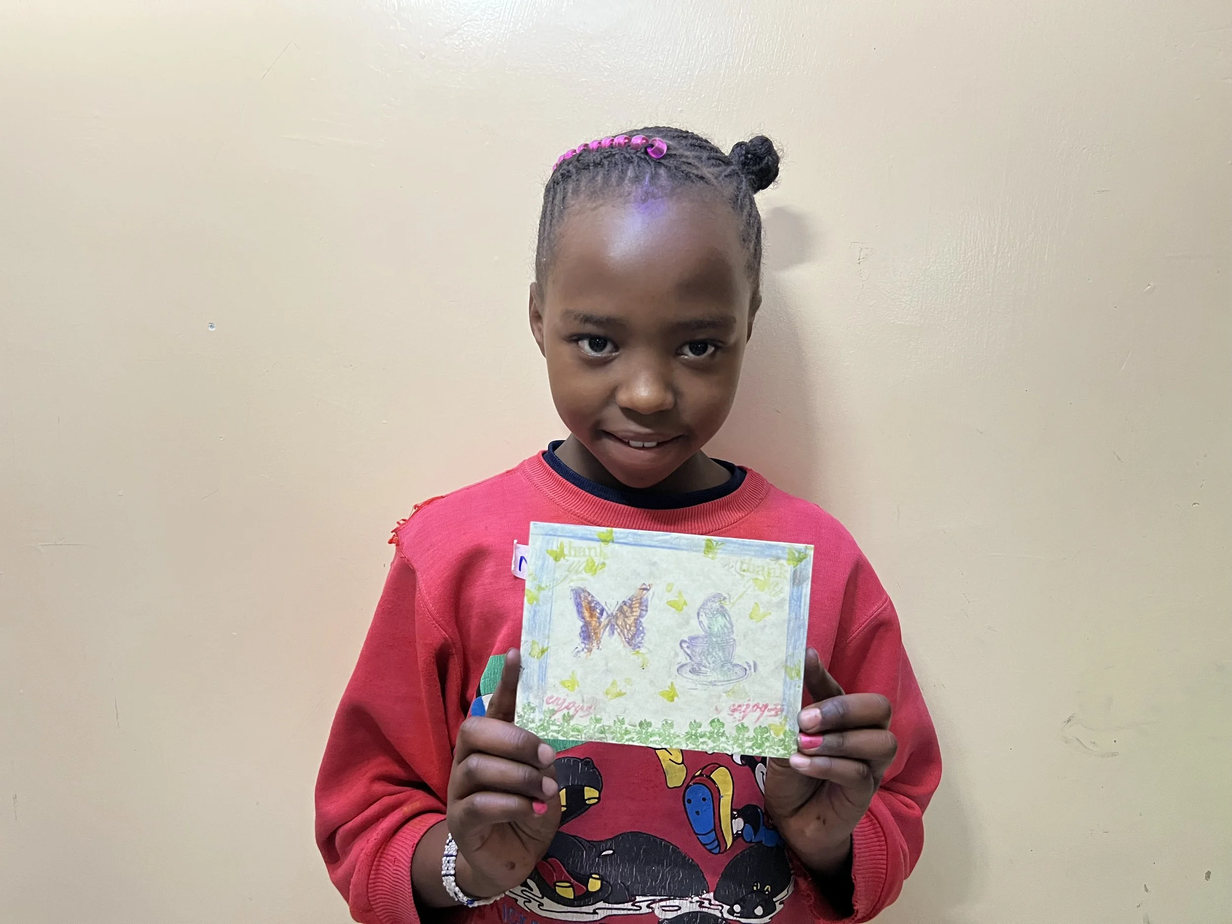 Young girl smiling and holding a handmade card with butterflies and leaves, standing against a light-colored wall.